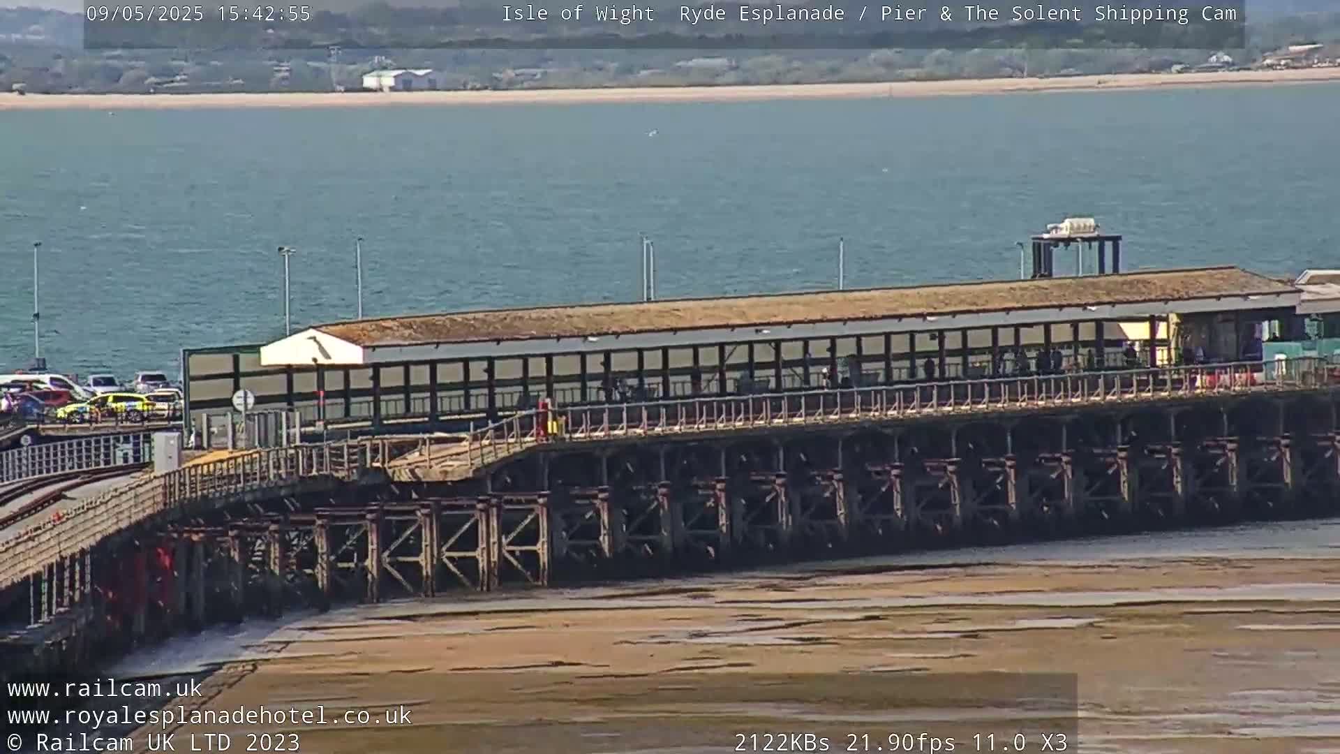 A long pier extends over calm water with a sandy shore visible at low tide under a sunny sky.