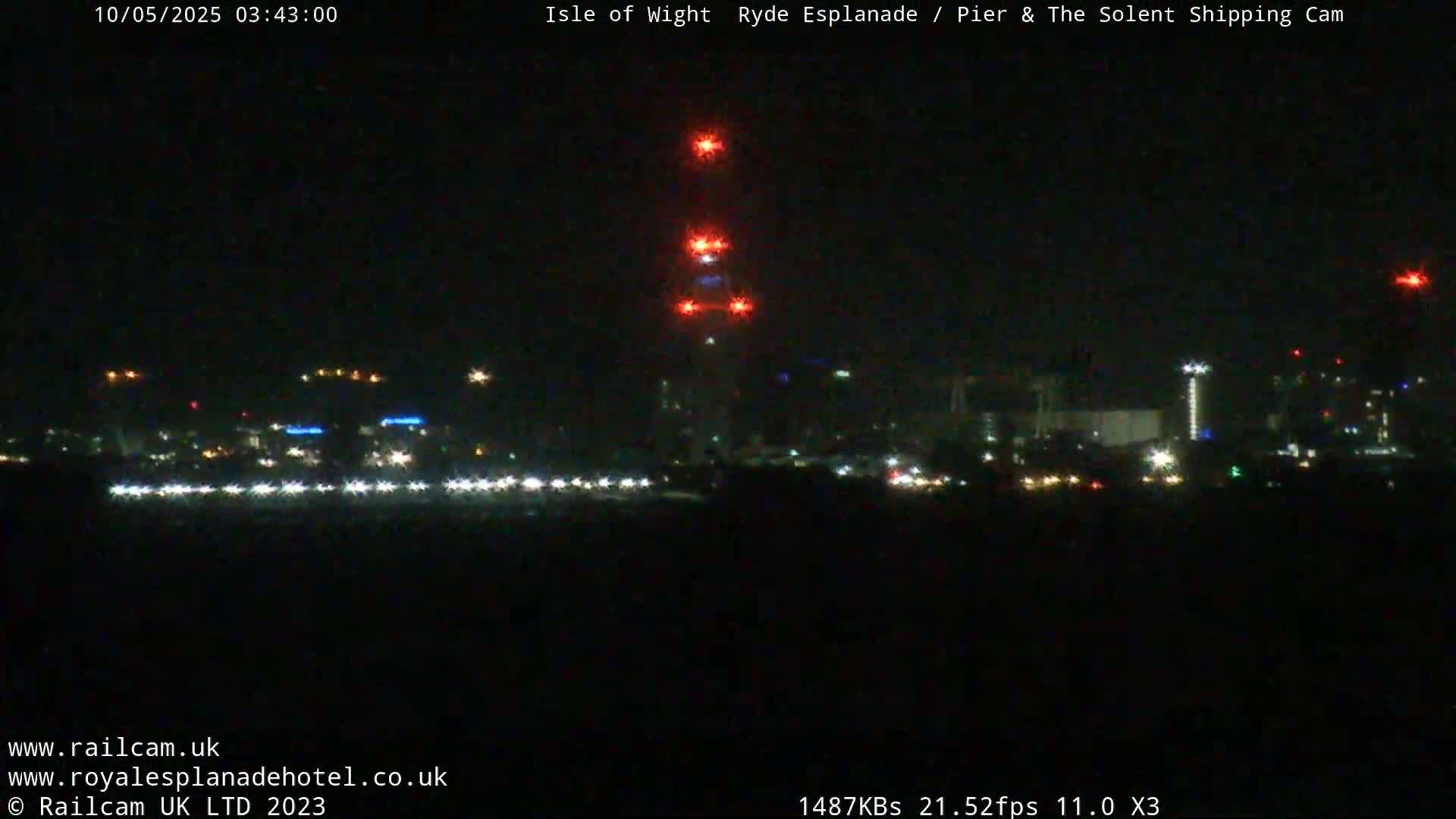 A nighttime long shot of a city skyline with a tall structure illuminated by red lights, seen across a dark body of water.