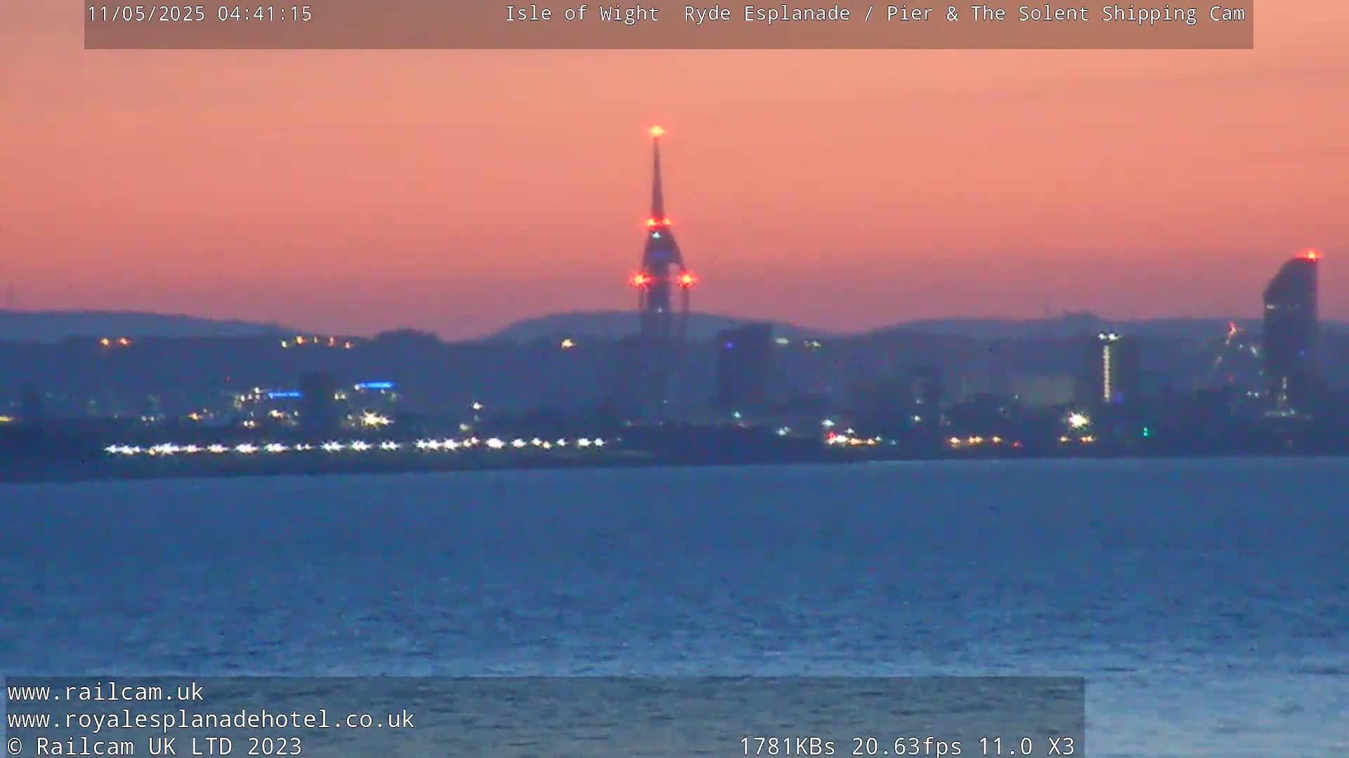 A calm body of water shows a city skyline at twilight, illuminated with lights against a pinkish-orange sky.