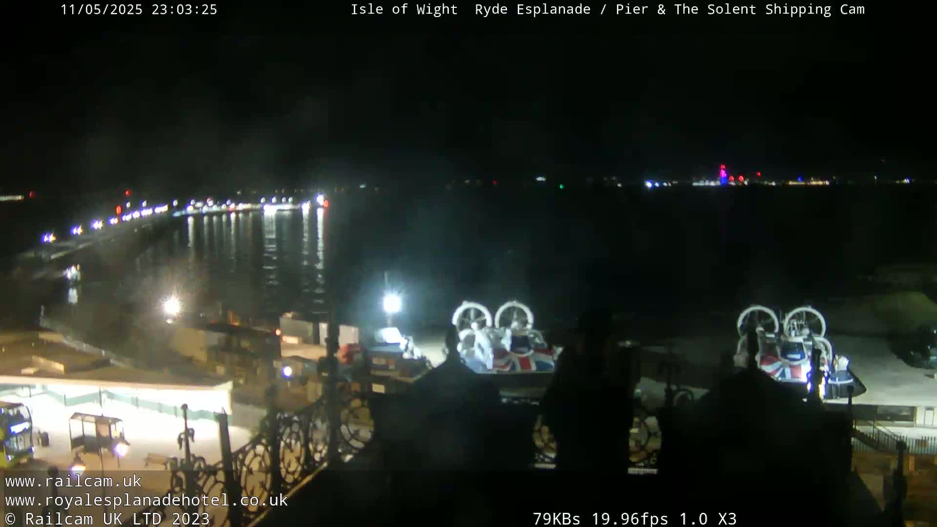 A nighttime view of a waterfront area with calm water, lights along a shoreline, and a hovercraft parked nearby.