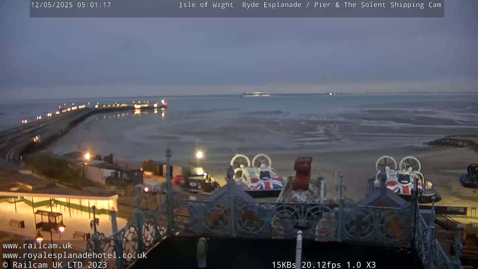 A low-light, pre-dawn view shows a beach with hovercrafts, a pier, and a body of water with a distant ship under a dusky sky.