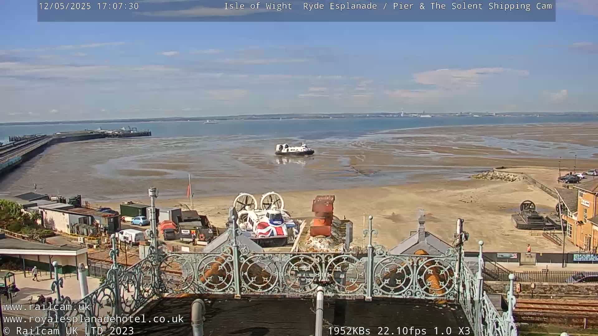 A hovercraft is on the water near a sandy beach, with other hovercrafts visible on land, under a partly sunny sky.