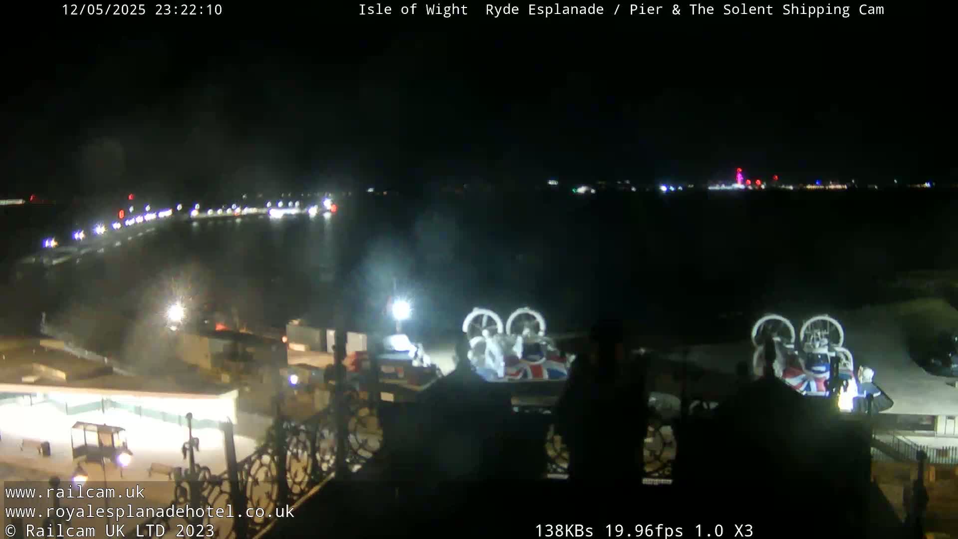A nighttime view of a waterfront area shows a body of water with lights across it and what appears to be a hovercraft with a Union Jack design parked on the shore.