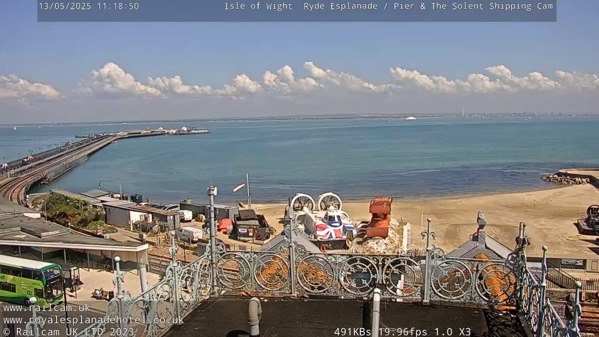 An elevated view shows a long pier extending into a calm body of water under a partly cloudy sky, with a hovercraft and other vehicles visible on the shore.