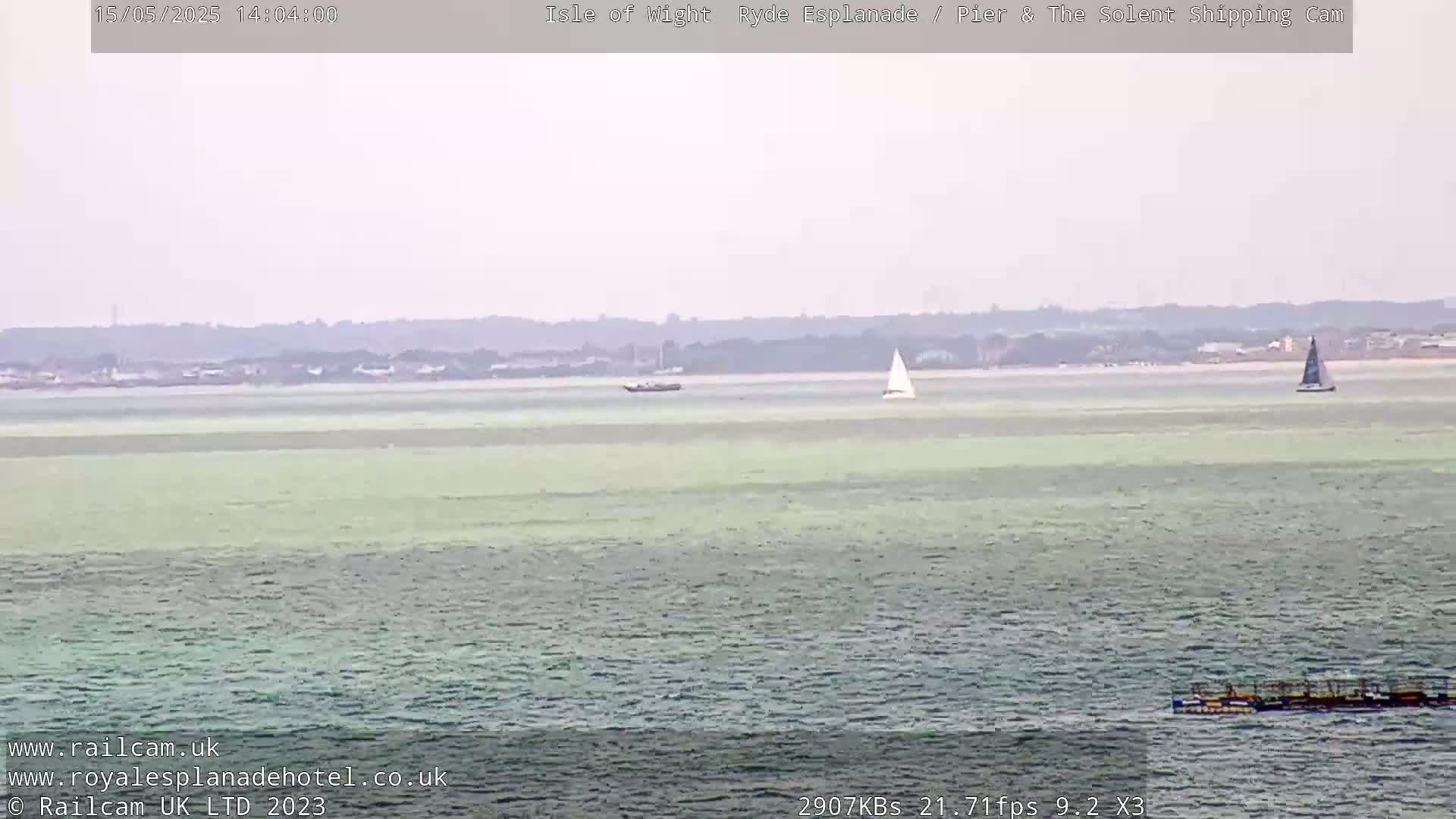 A calm, hazy day shows a wide expanse of greenish-grey water with two sailboats and a distant shoreline.
