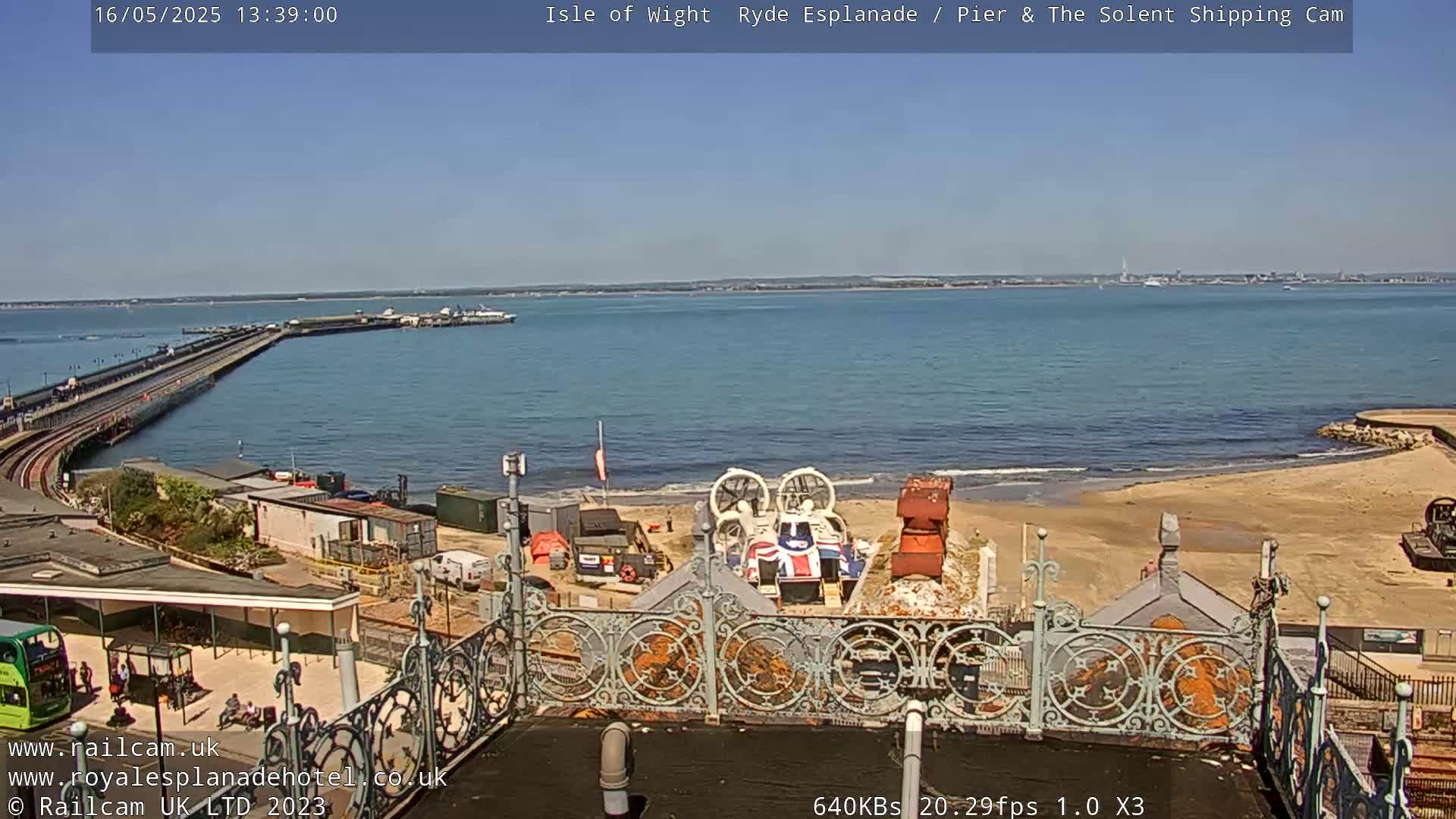 A hovercraft sits on a sandy beach near a pier and calm blue water under a clear blue sky.