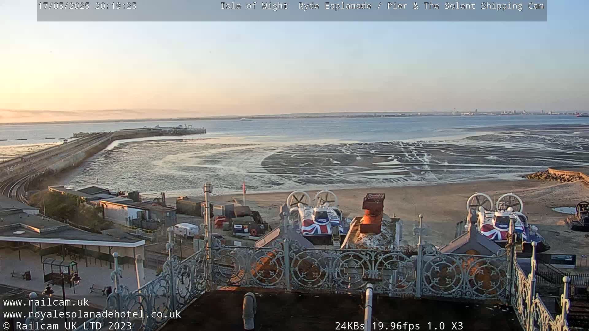 An expansive coastal view at sunset shows a pier extending into a wide expanse of mudflats, with two hovercrafts parked on the shore in the foreground.