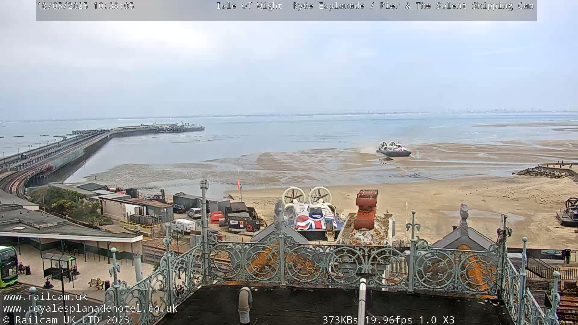 An overcast day shows a beach with two hovercrafts, a long pier extending into the water, and a building with ornate metal railings in the foreground.
