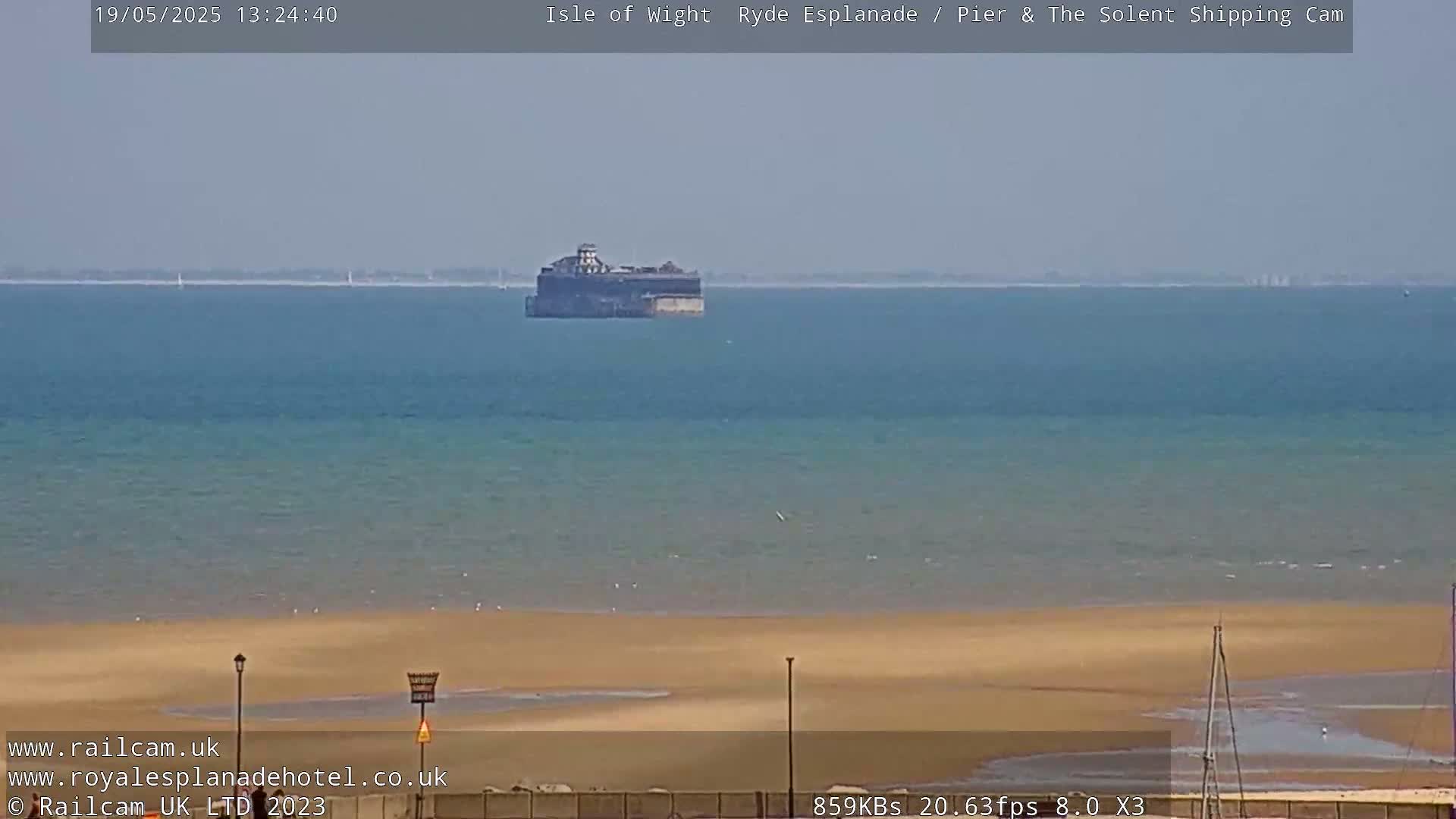 A wide shot of a sandy beach and calm ocean on a sunny day, with a distant, dark structure visible in the water.