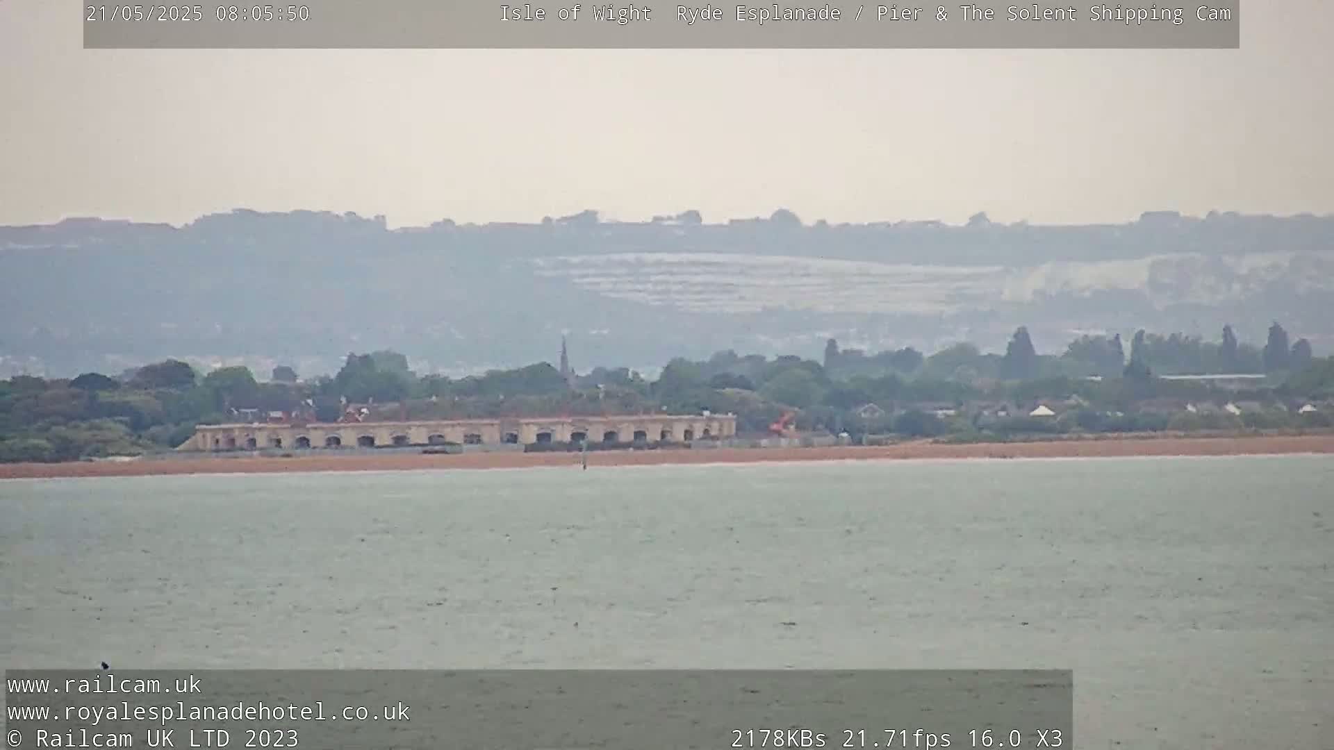 A hazy view of a calm body of water shows a long, low building near a sandy shore, with distant hills and trees under an overcast sky.