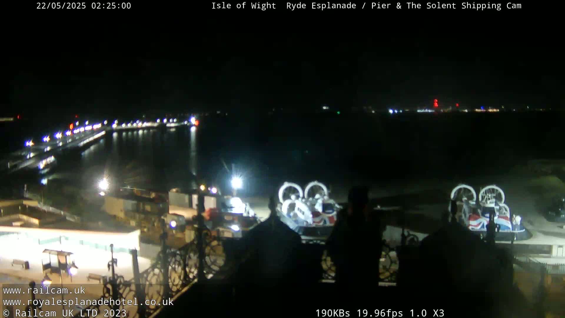 A nighttime, seaside view shows a pier with lights, a body of water, and two hovercrafts parked near a building.
