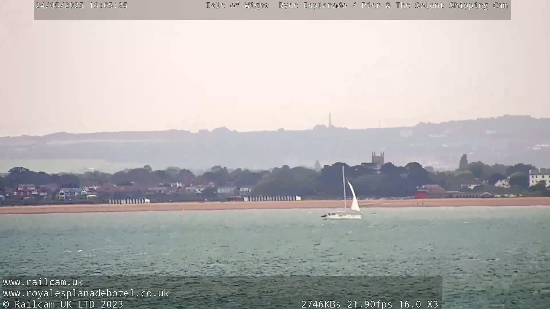 A sailboat is visible on calm, gray water in front of a hazy, distant shoreline with buildings and trees under a muted sky.