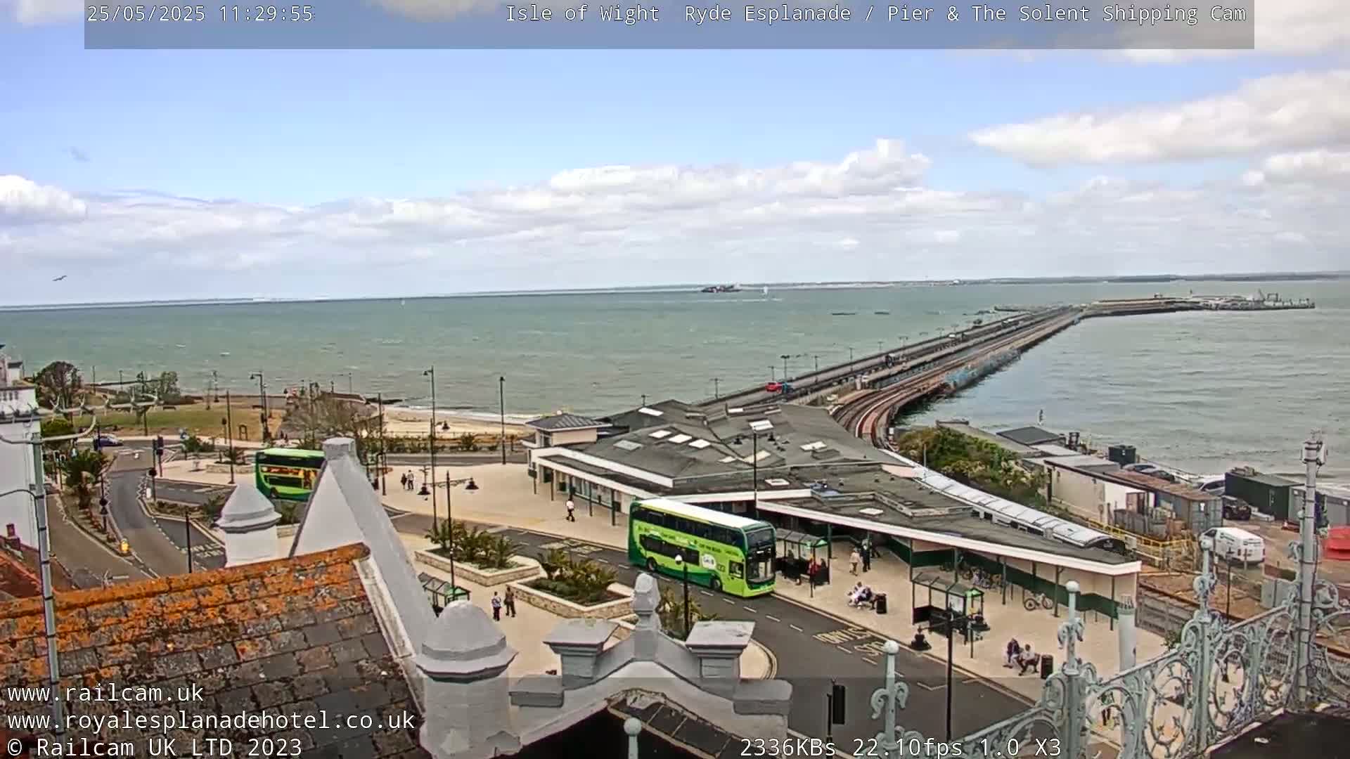 An elevated view of a pier extending into a calm sea under a partly cloudy sky, with buses and people visible near a station on land.
