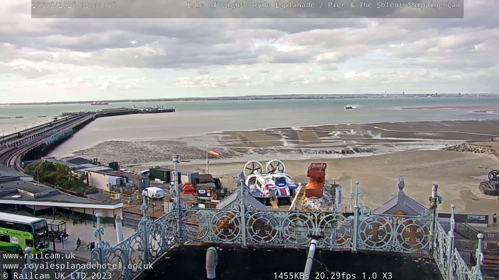 An overcast day shows a pier extending into a calm body of water with exposed sandbars at low tide, a hovercraft is docked near a building with an ornate railing, and a bus is parked near the building.