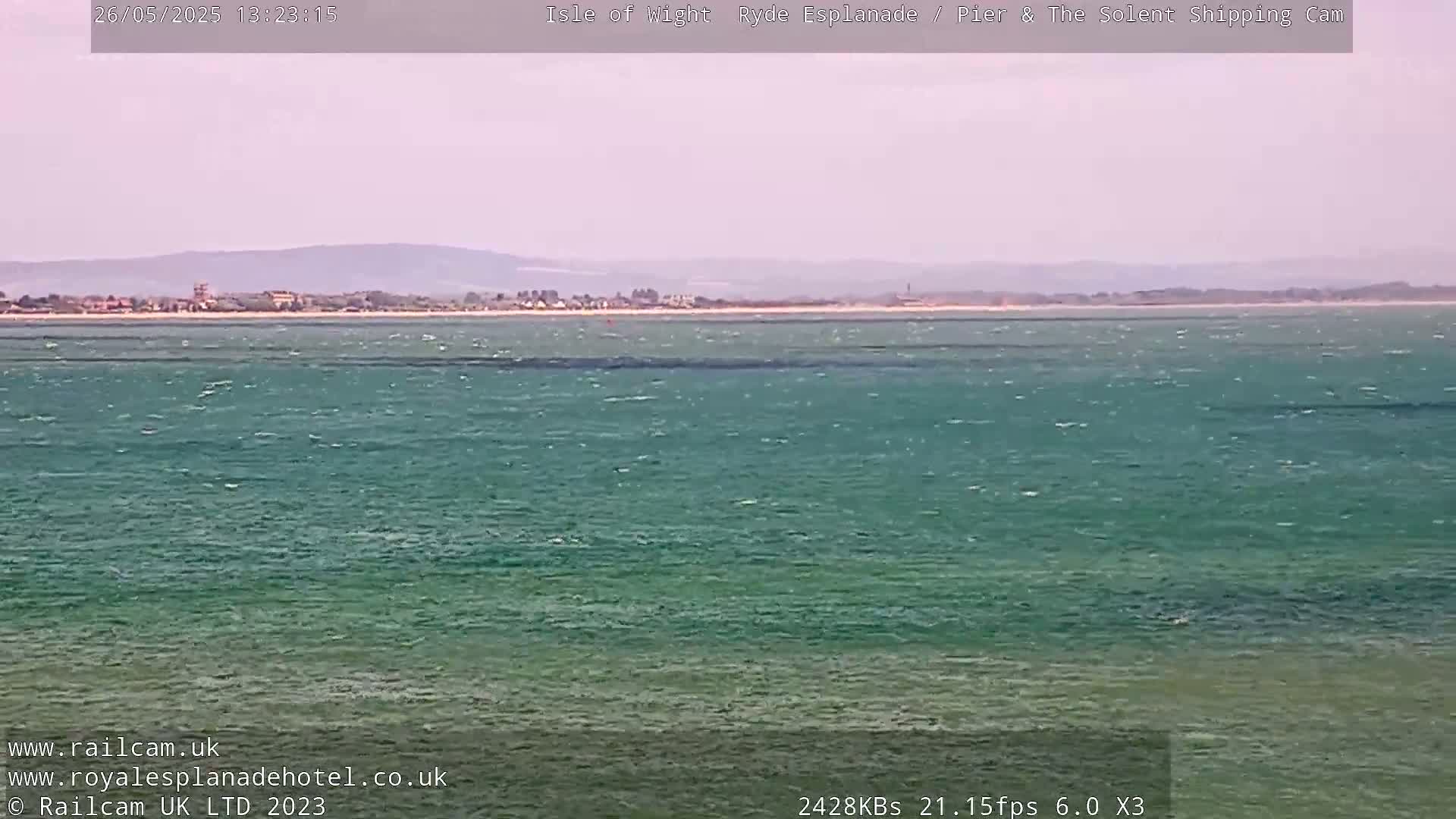 A wide shot shows a calm body of teal-green water stretching to a distant shoreline under a hazy sky.