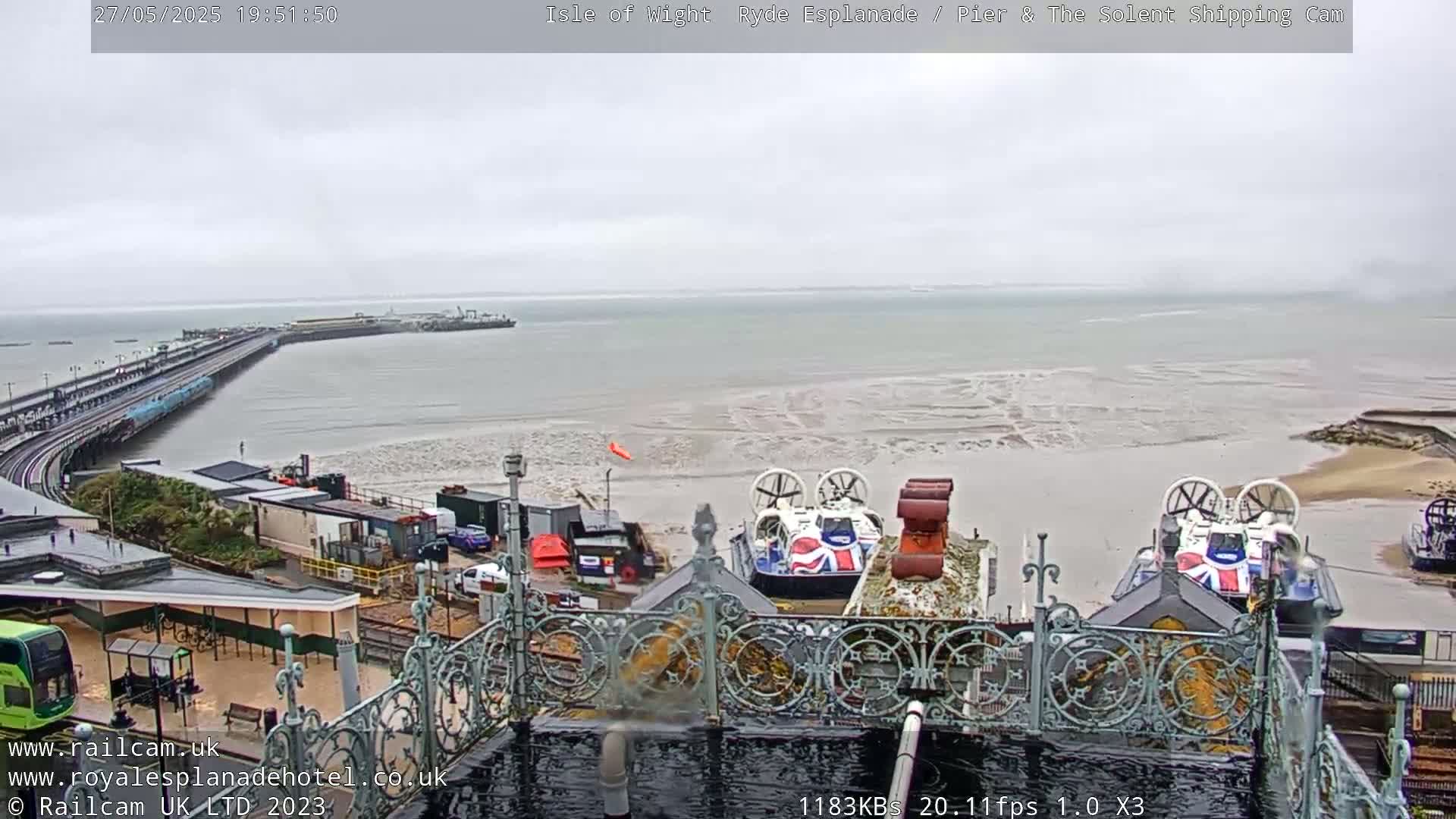 An overcast day reveals a pier extending into a body of water with exposed mudflats, two hovercrafts near the shore, and a view from an elevated vantage point overlooking a town.