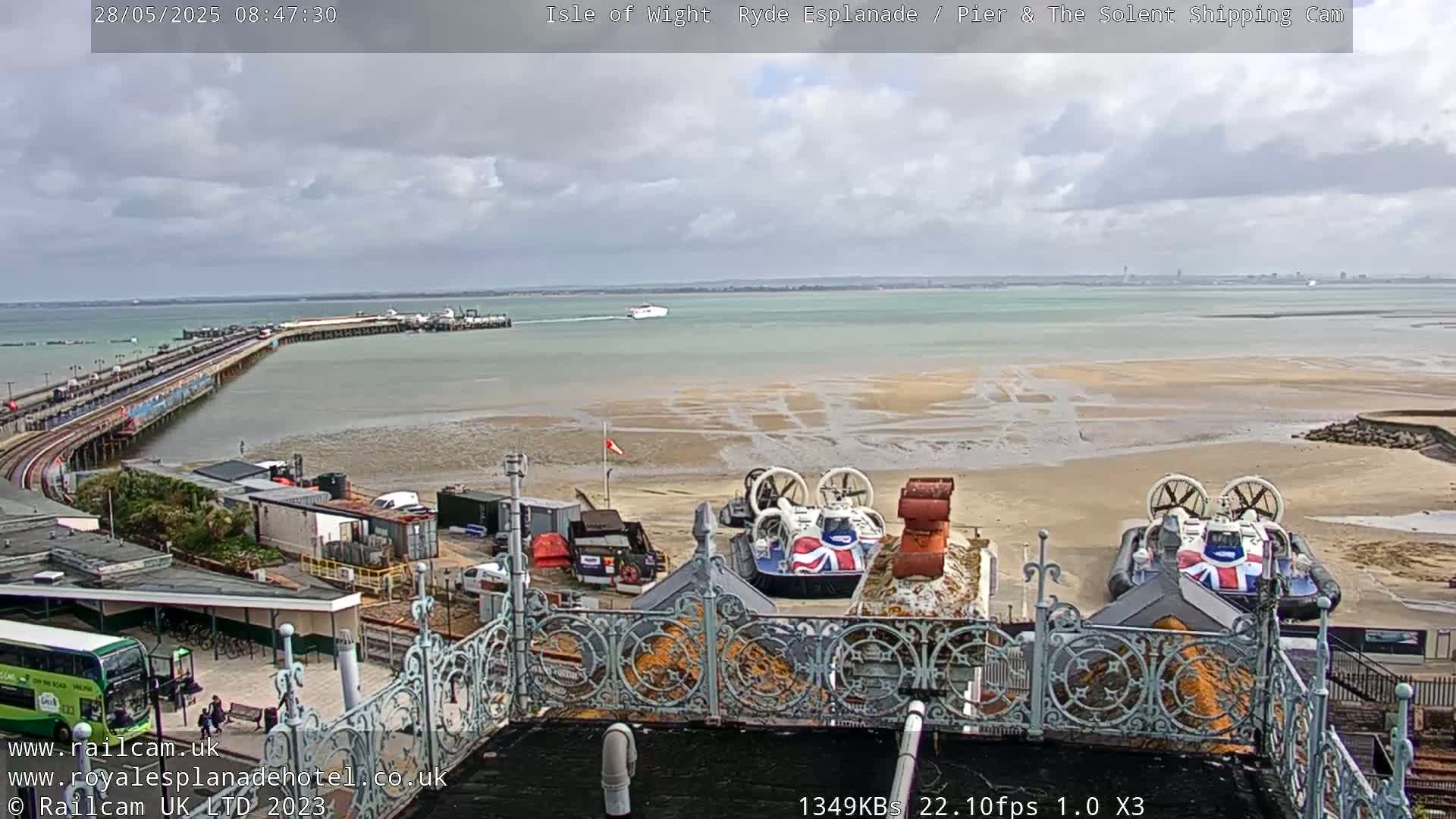 An overcast day shows a pier extending into the water, two hovercrafts on the shore, and a ferry in the distance.