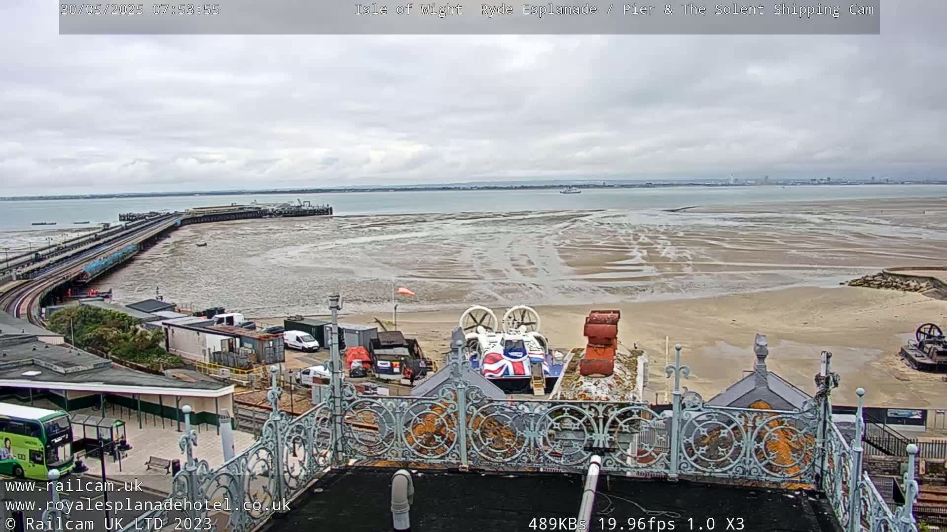 An overcast day reveals a view of a pier, a hovercraft, and a wide expanse of tidal mudflats leading to calm water and a distant shoreline.
