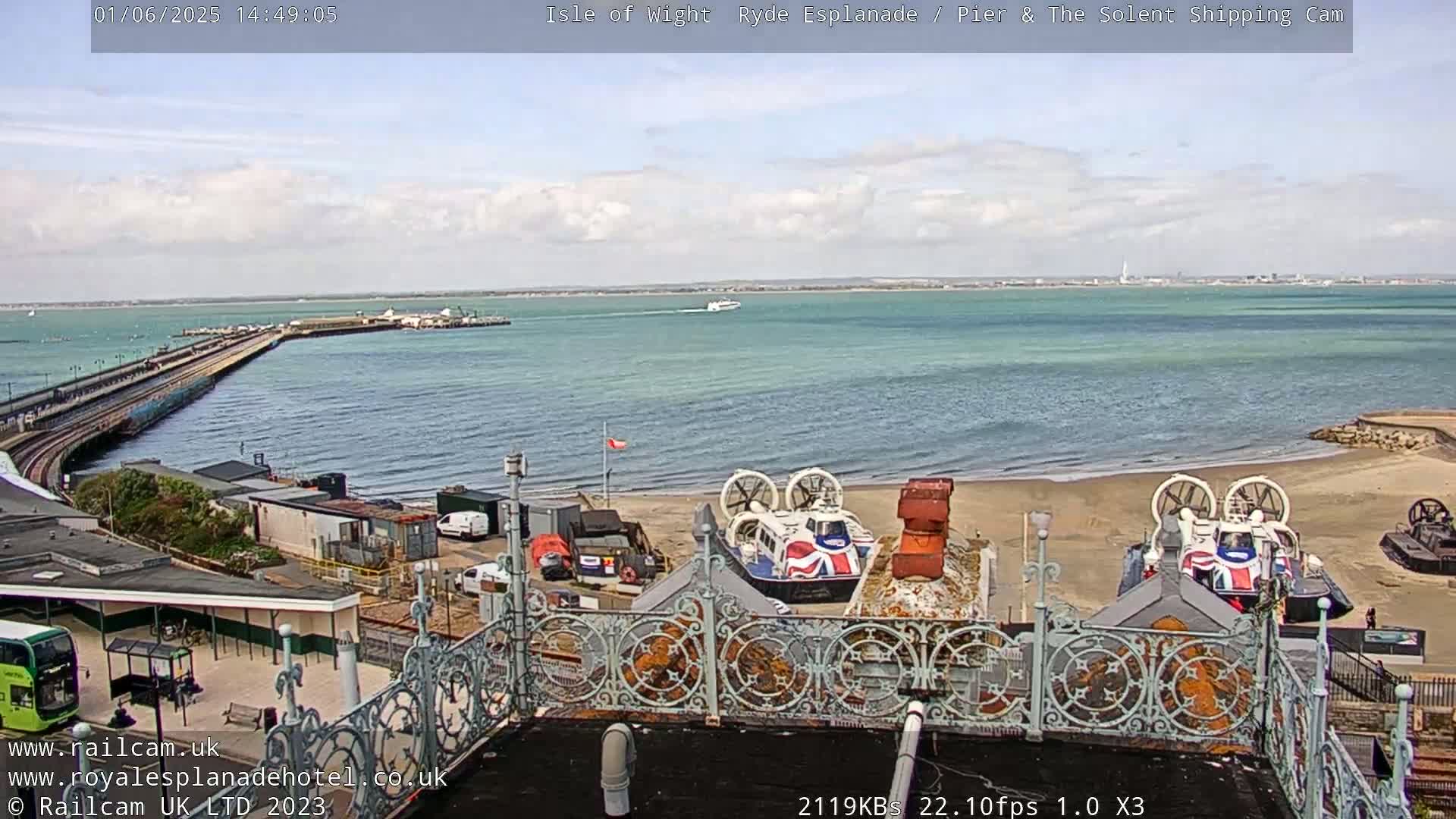 A partly cloudy day shows a seaside scene with a pier extending into the water, several hovercrafts on a sandy beach, and a body of water stretching to a distant shoreline.