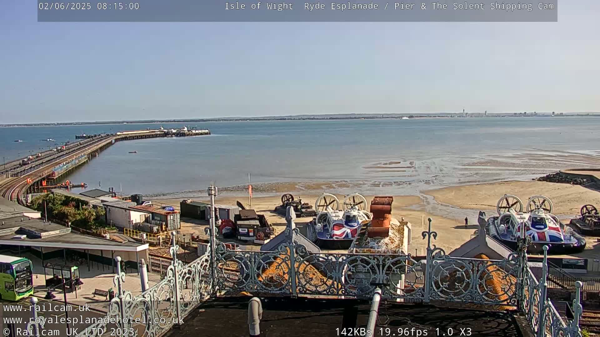 A sunny day overlooking a pier, a sandy beach with hovercrafts, and a calm body of water extending to a distant shoreline.