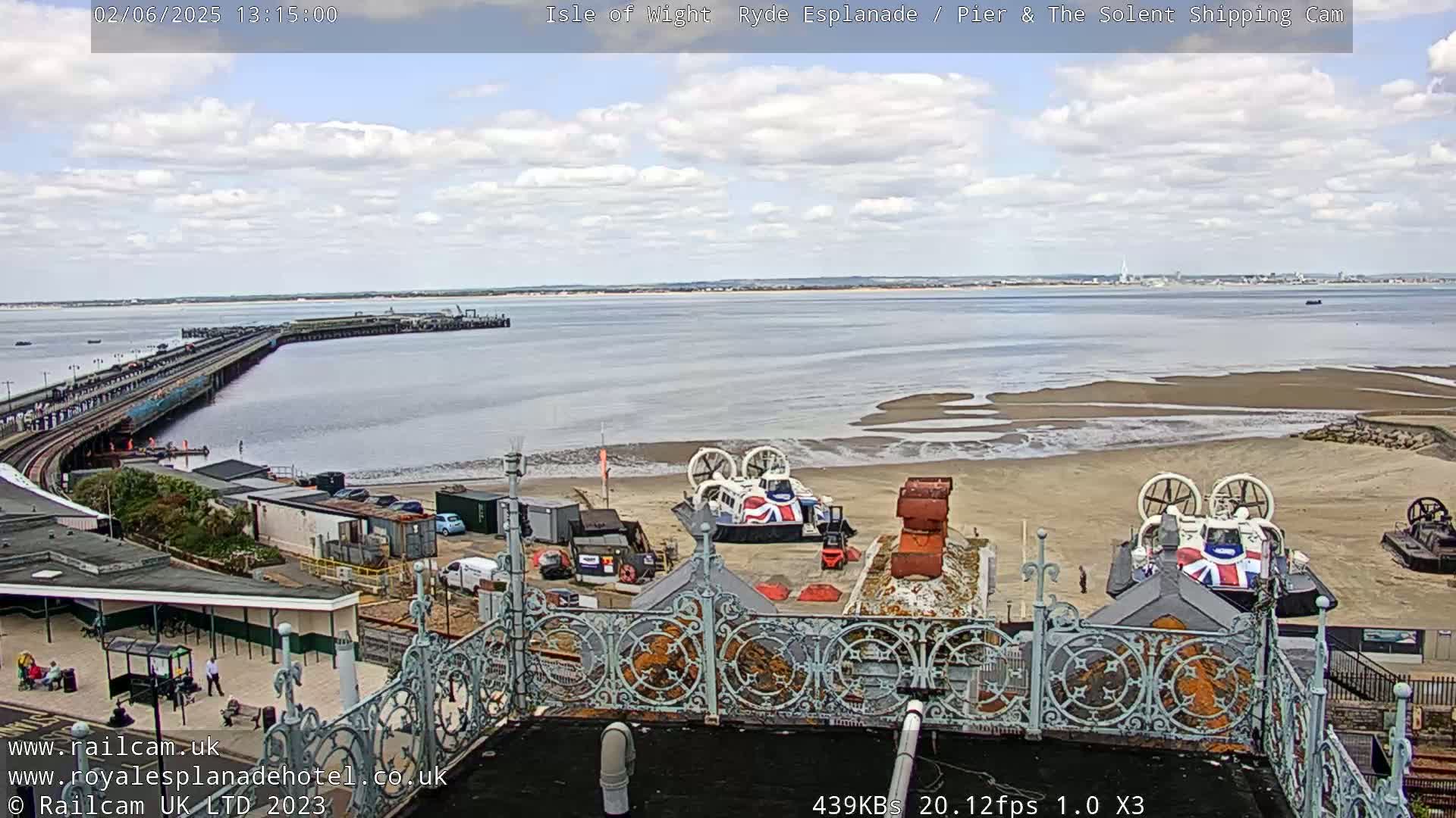 A partly cloudy day shows a pier extending into a body of water with a sandy beach, several hovercrafts, and buildings.