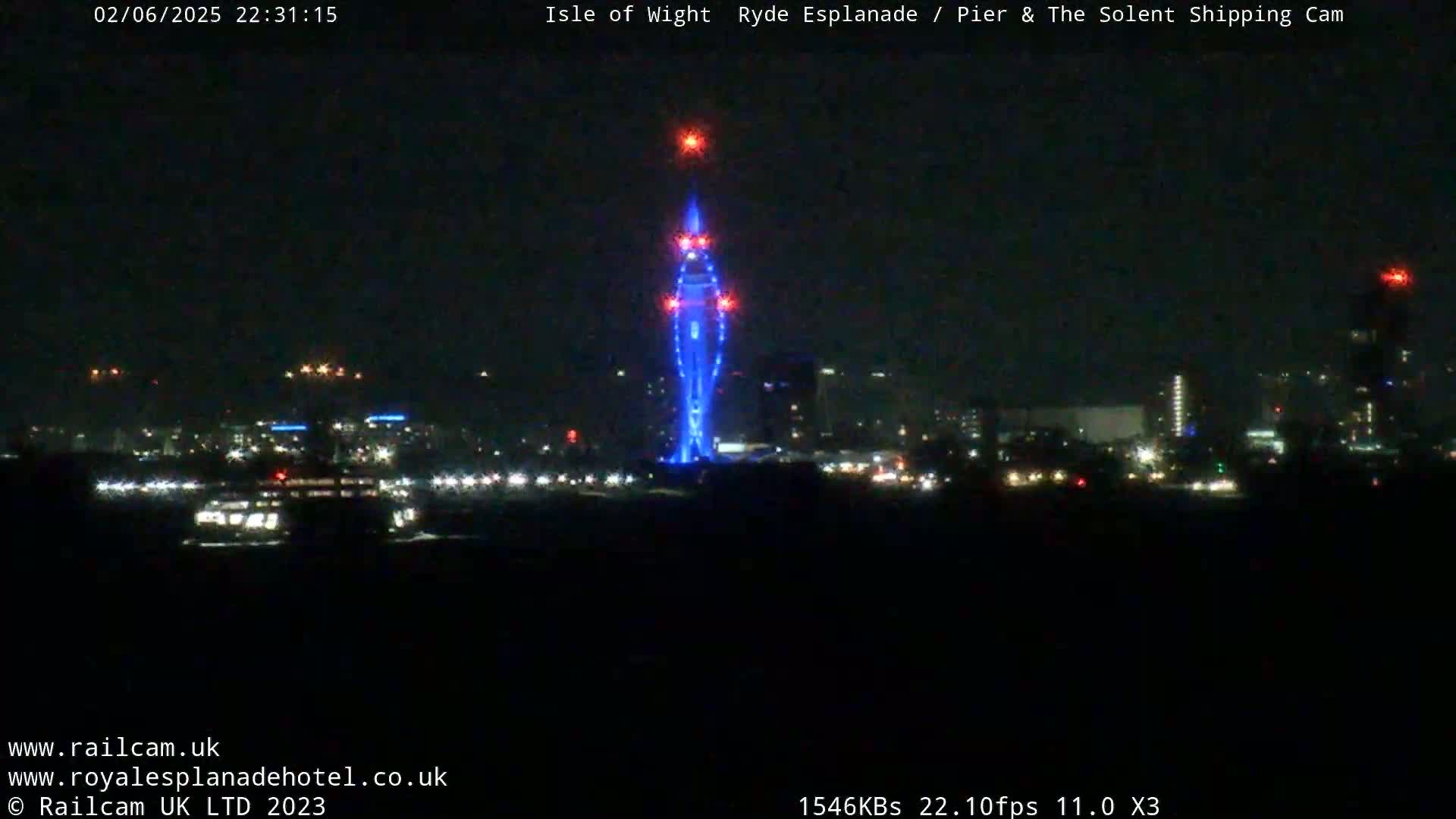 A nighttime long shot shows a city skyline across water, with a tall structure illuminated in blue and red lights in the center.