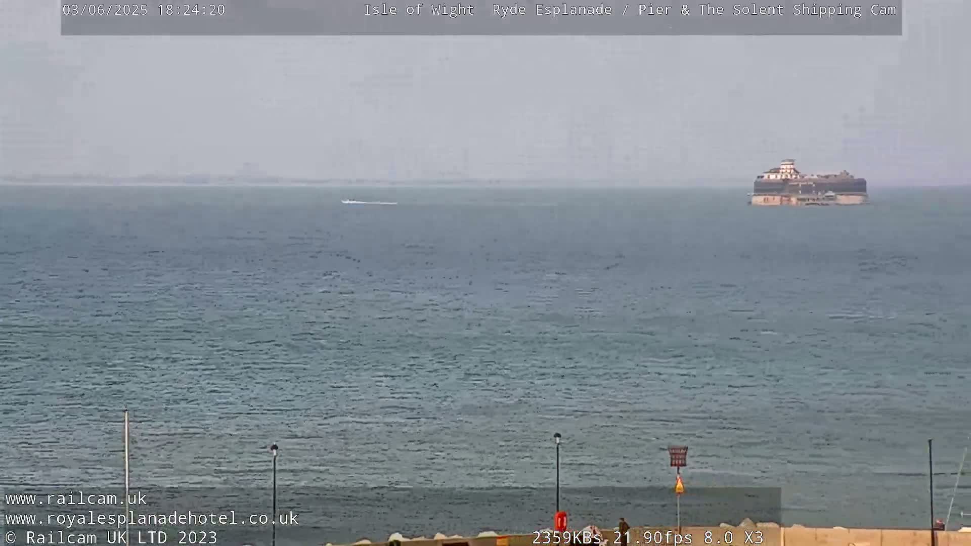 A calm, gray day shows a body of water with a distant structure and a small boat visible in the far distance.