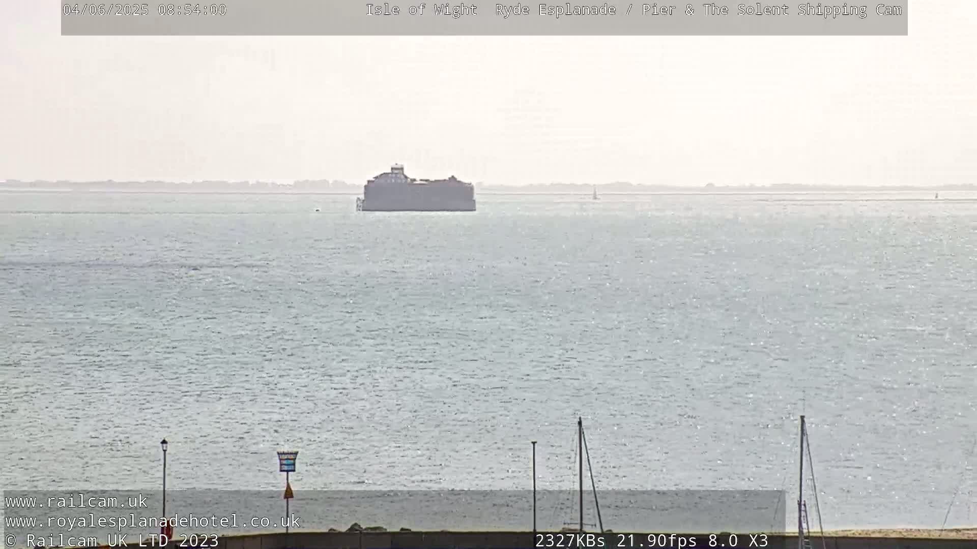 A hazy, calm sea is shown with a large, dark structure in the mid-ground and a distant shoreline.