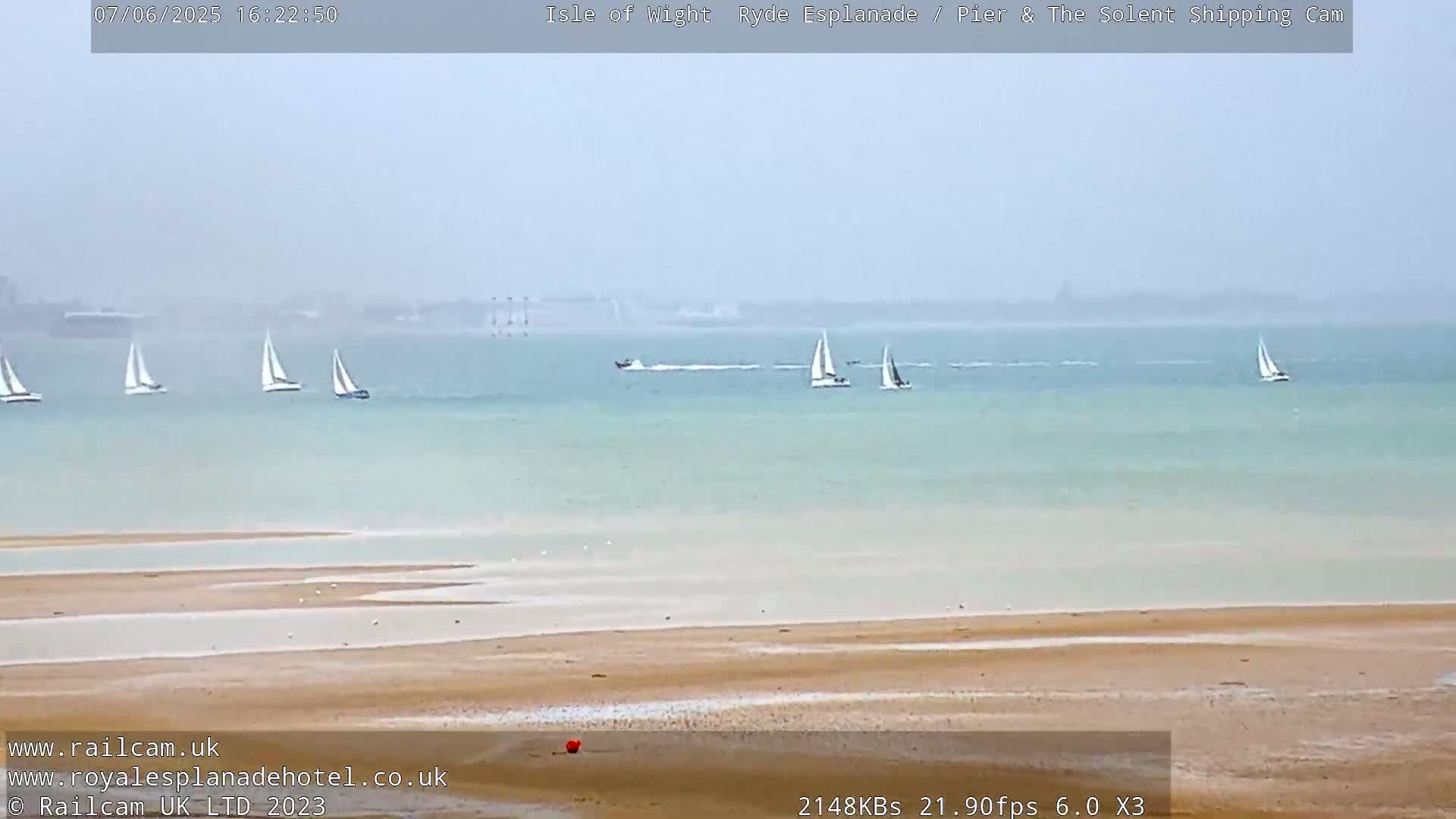 Several sailboats are visible on a calm, light-grey body of water, with a sandy beach in the foreground.