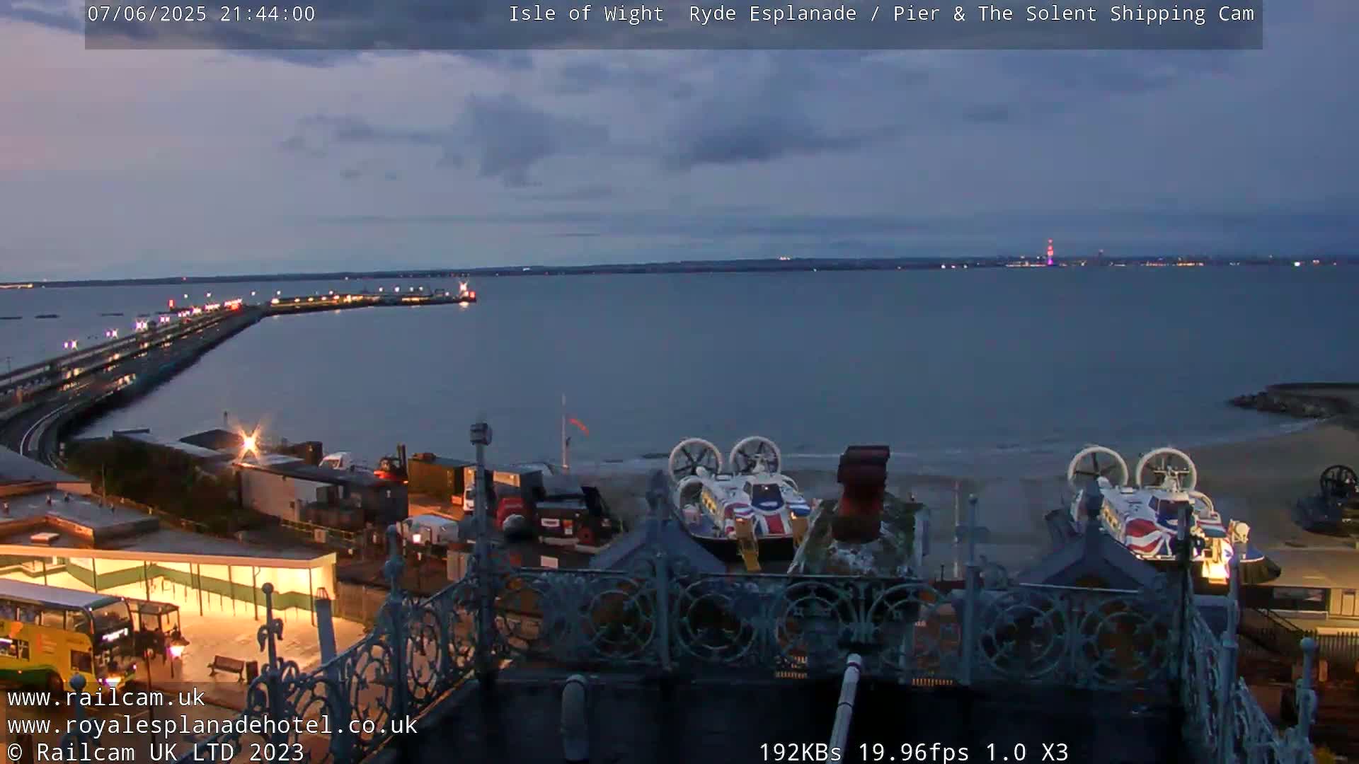 A twilight view of a pier extending into a calm body of water, with hovercrafts parked on a nearby beach.