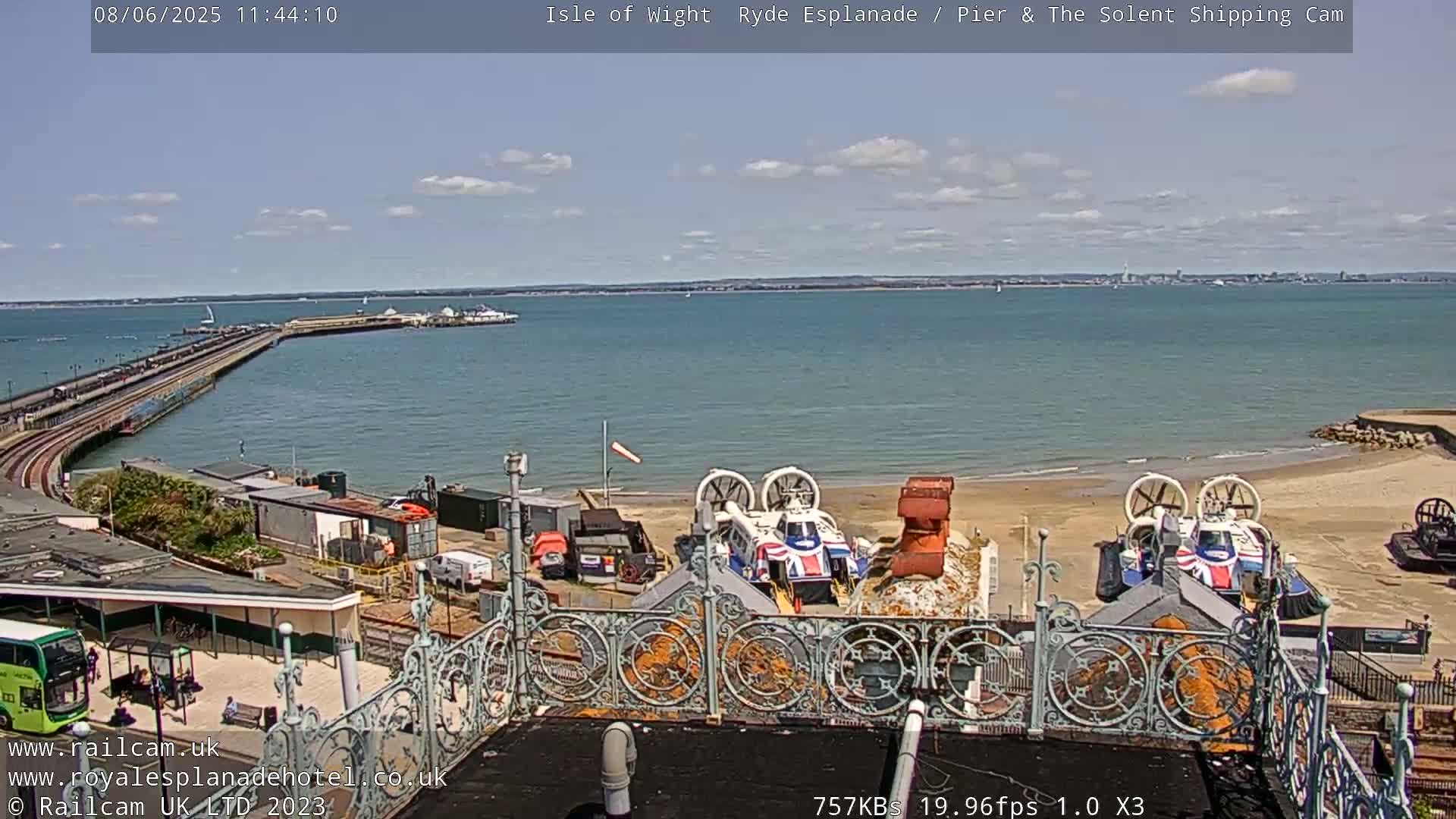 A seaside view on a sunny day shows a long pier extending into the water, hovercrafts on a sandy beach, and a distant cityscape across the bay.