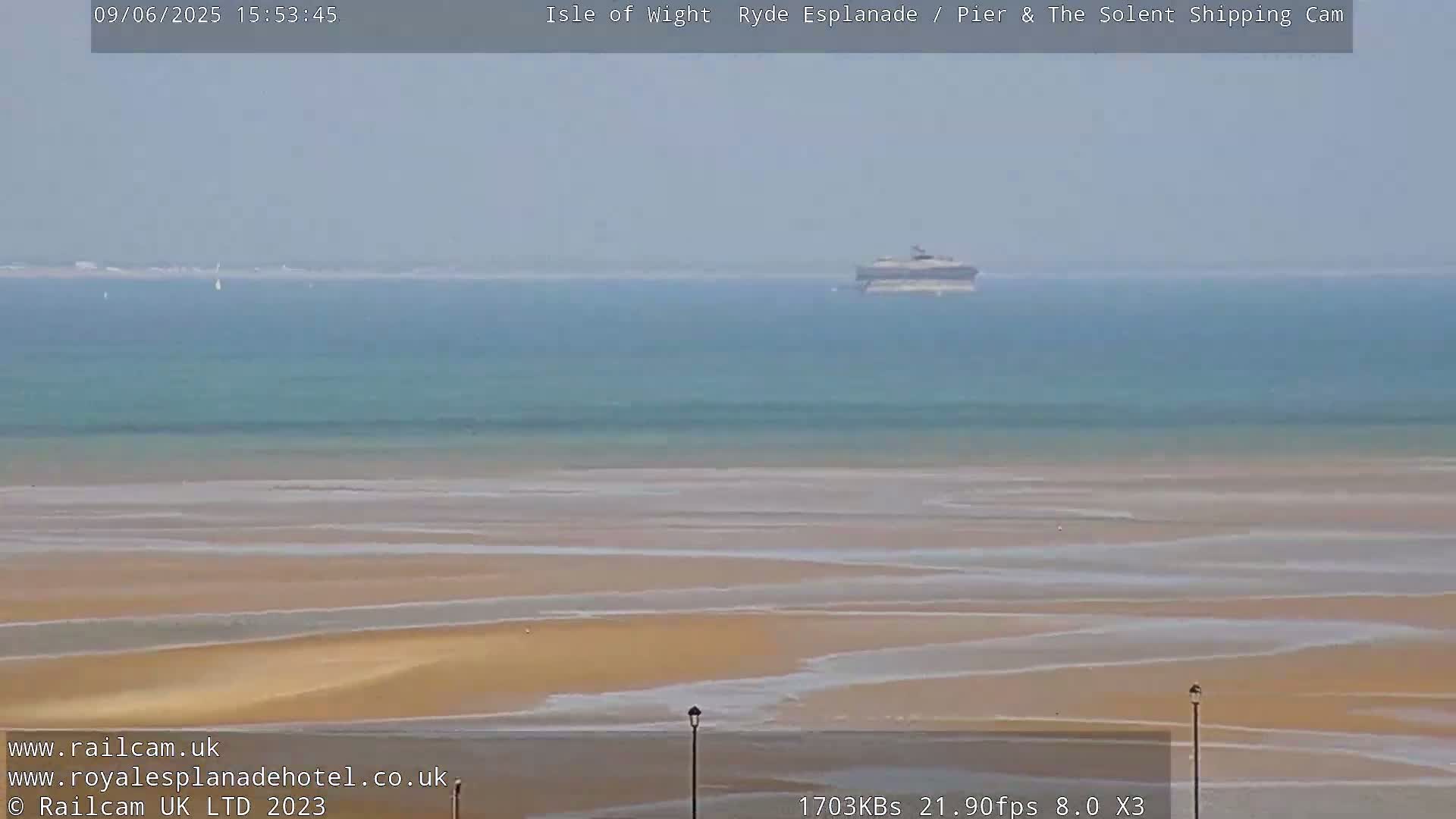 A hazy, sunny day shows a large body of water with a ship in the distance, a wide expanse of light brown sand at low tide, and two lampposts in the foreground.