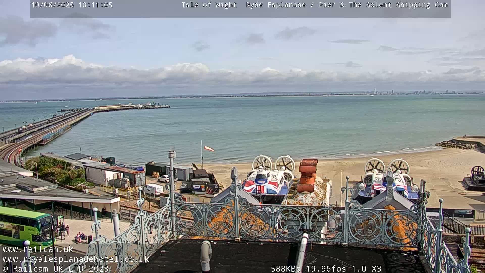 A partly cloudy day shows a long pier extending into a calm body of water, with two hovercrafts parked on a sandy beach nearby.