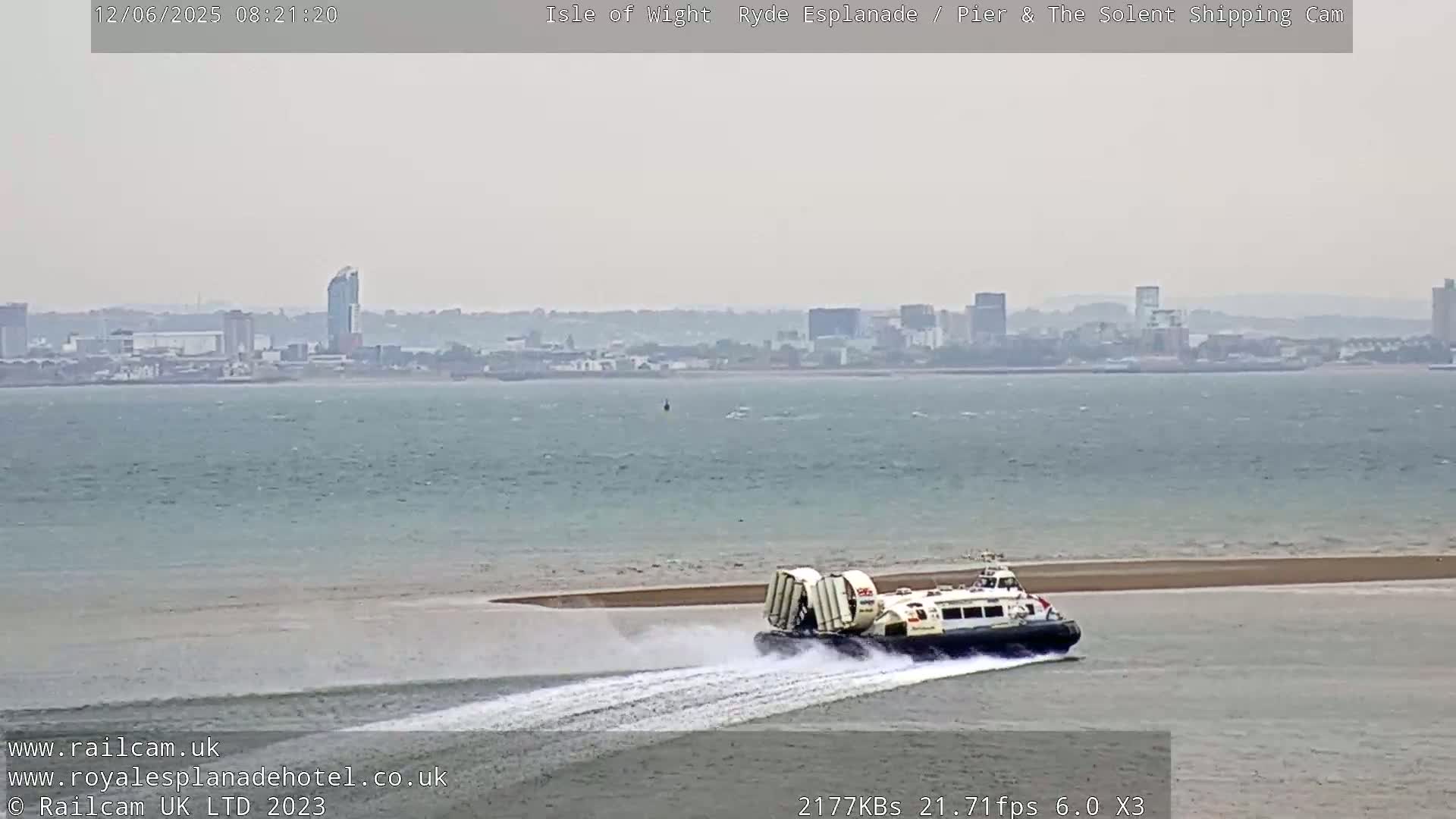 A hovercraft moves across the water toward a shoreline, with a hazy cityscape visible in the distance under an overcast sky.
