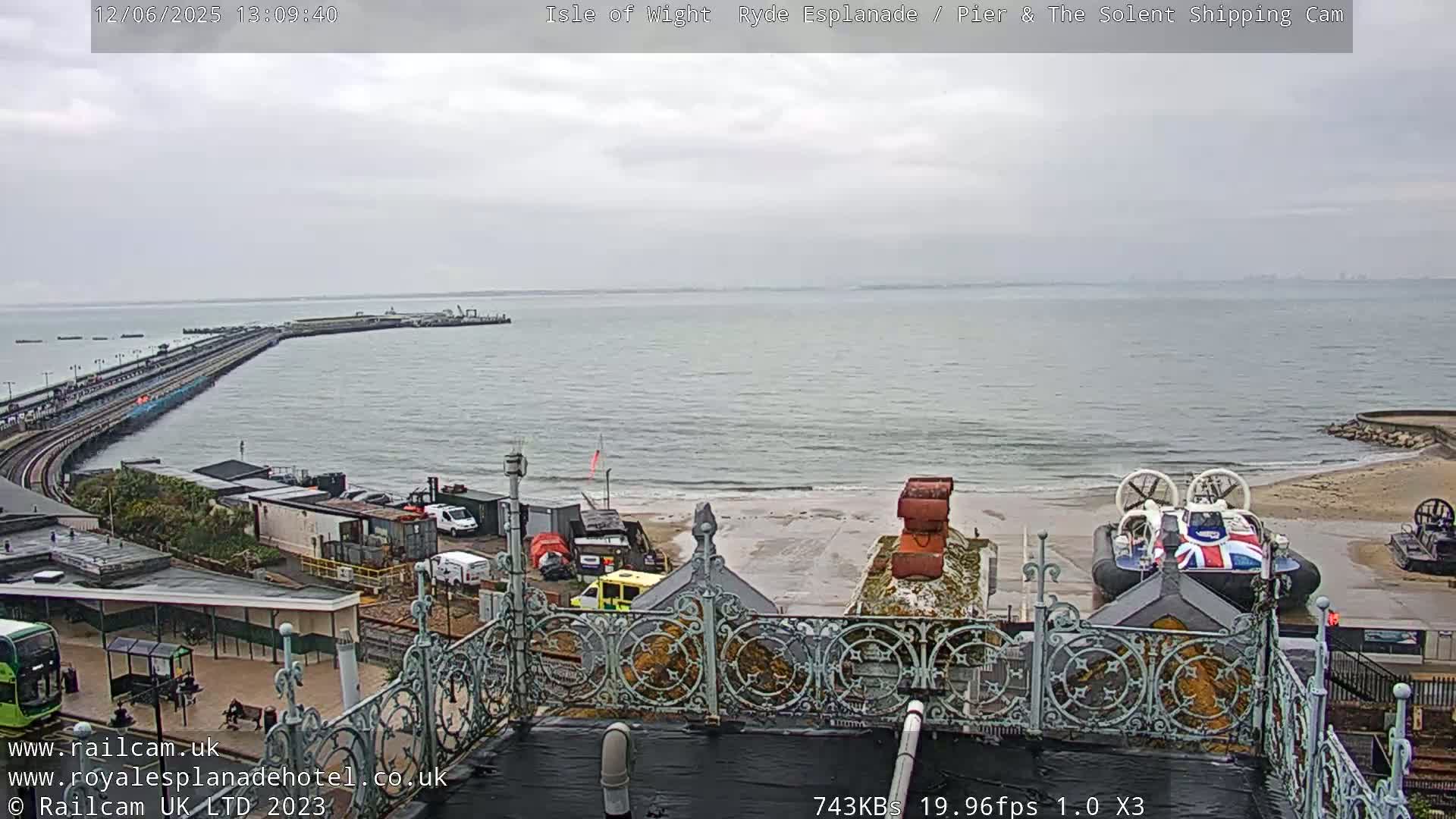 A hovercraft sits on a beach near a long pier extending into an overcast ocean.