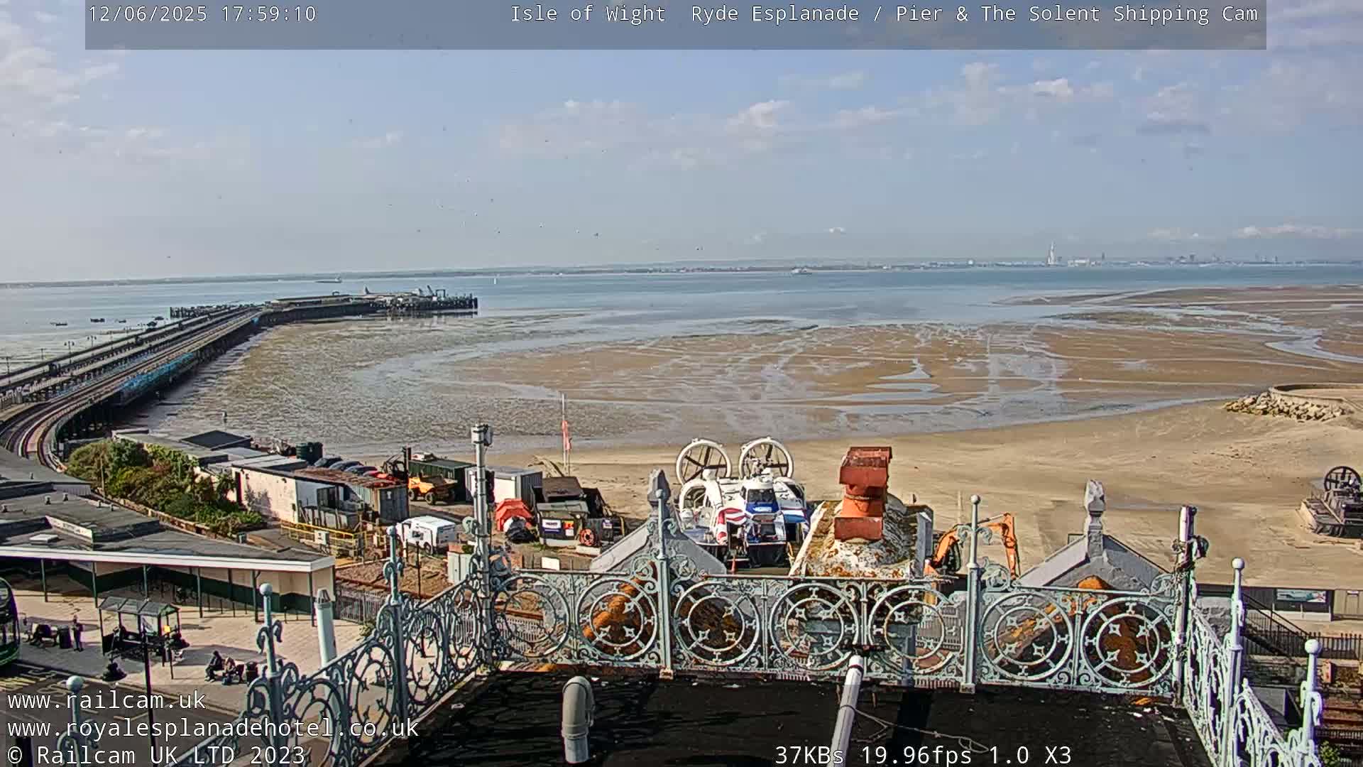 A hovercraft sits on a sandy beach near a pier and train tracks, under a mostly sunny sky.