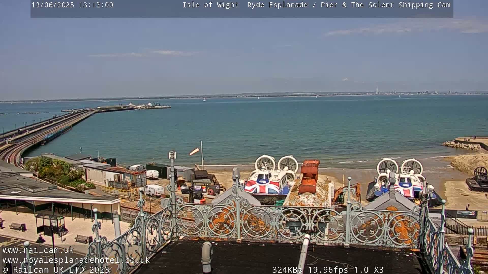 A sunny day overlooking a pier extending into a calm body of water, with two hovercrafts on a sandy beach in the foreground.