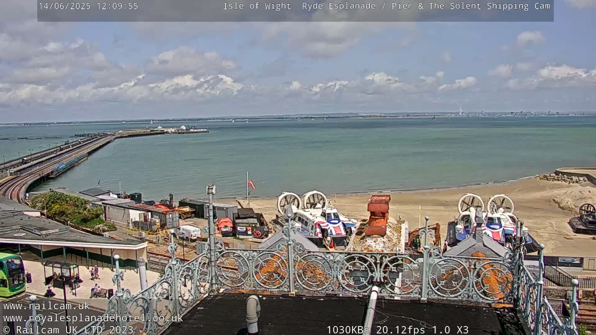A partly cloudy day overlooking a pier, calm water, a sandy beach with hovercrafts, and a distant cityscape.