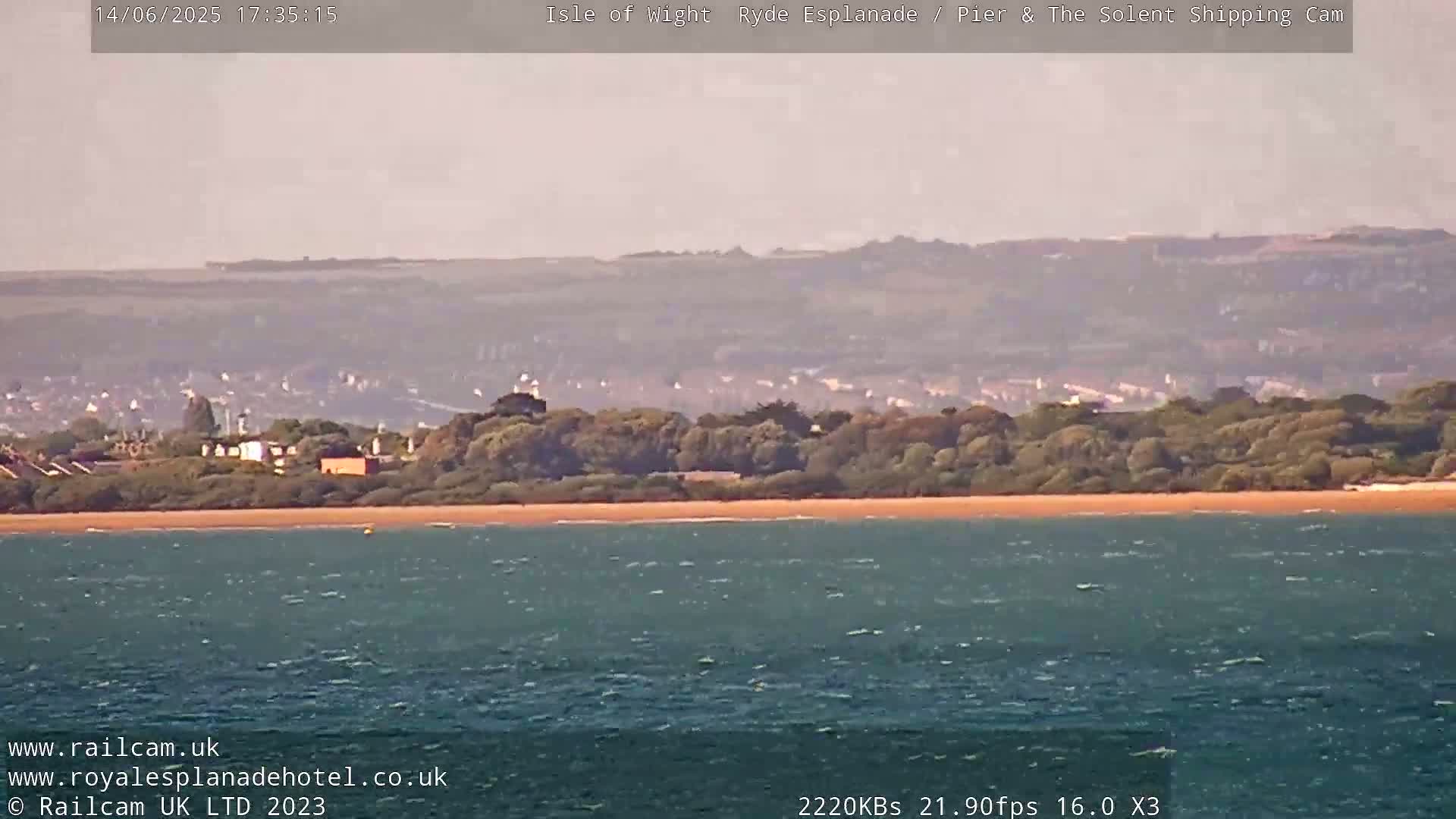 A hazy, calm day reveals a sandy beach backed by trees and distant buildings, all overlooking a body of water.