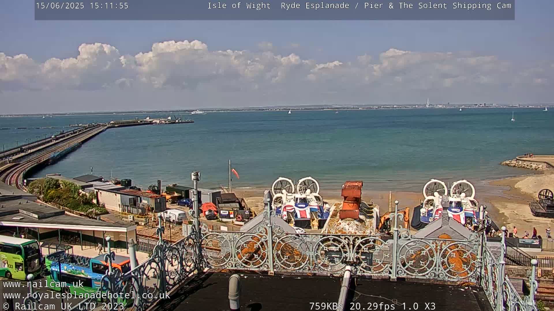 A partly cloudy day shows a pier extending into a body of water with hovercrafts on the shore, viewed from a high vantage point.