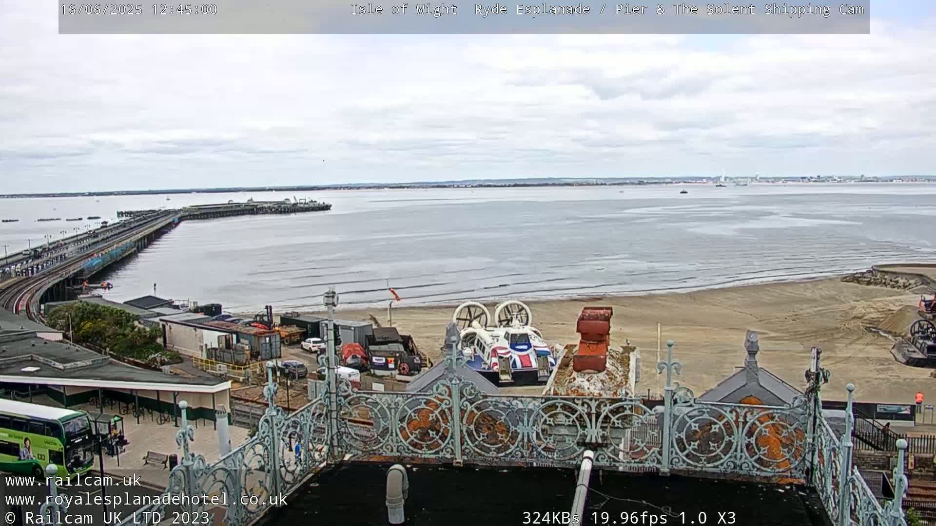 An overcast day shows a long pier extending into a calm body of water, with a hovercraft and beach visible in the foreground, all seen from an elevated vantage point.