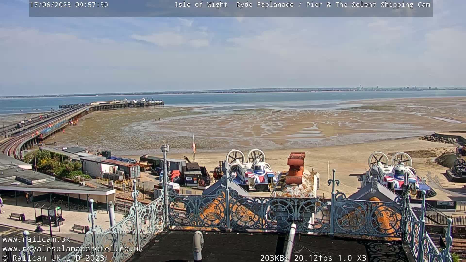 A mostly sunny day overlooks a pier, a sandy beach with hovercrafts, and a calm body of water extending to a distant shoreline.