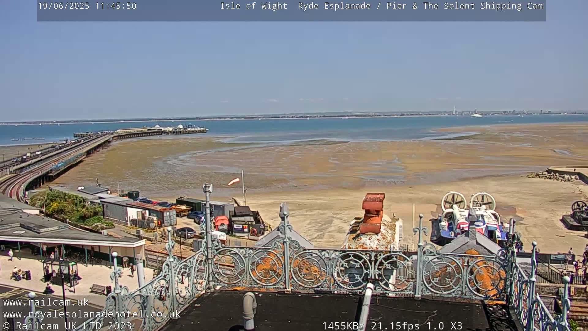 A sunny view from a rooftop shows a long pier extending into a calm body of water, with a sandy beach and hovercrafts visible in the foreground.