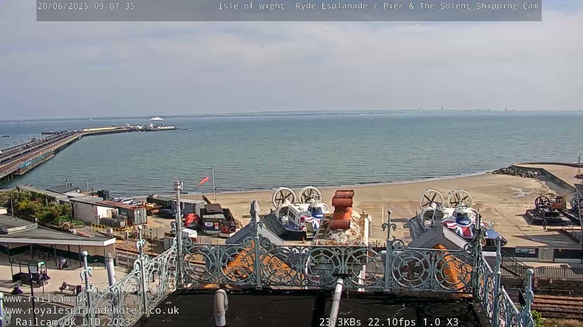 A sunny view of a beach with two hovercrafts parked on the sand, a pier extending into calm water, and a distant ferry.