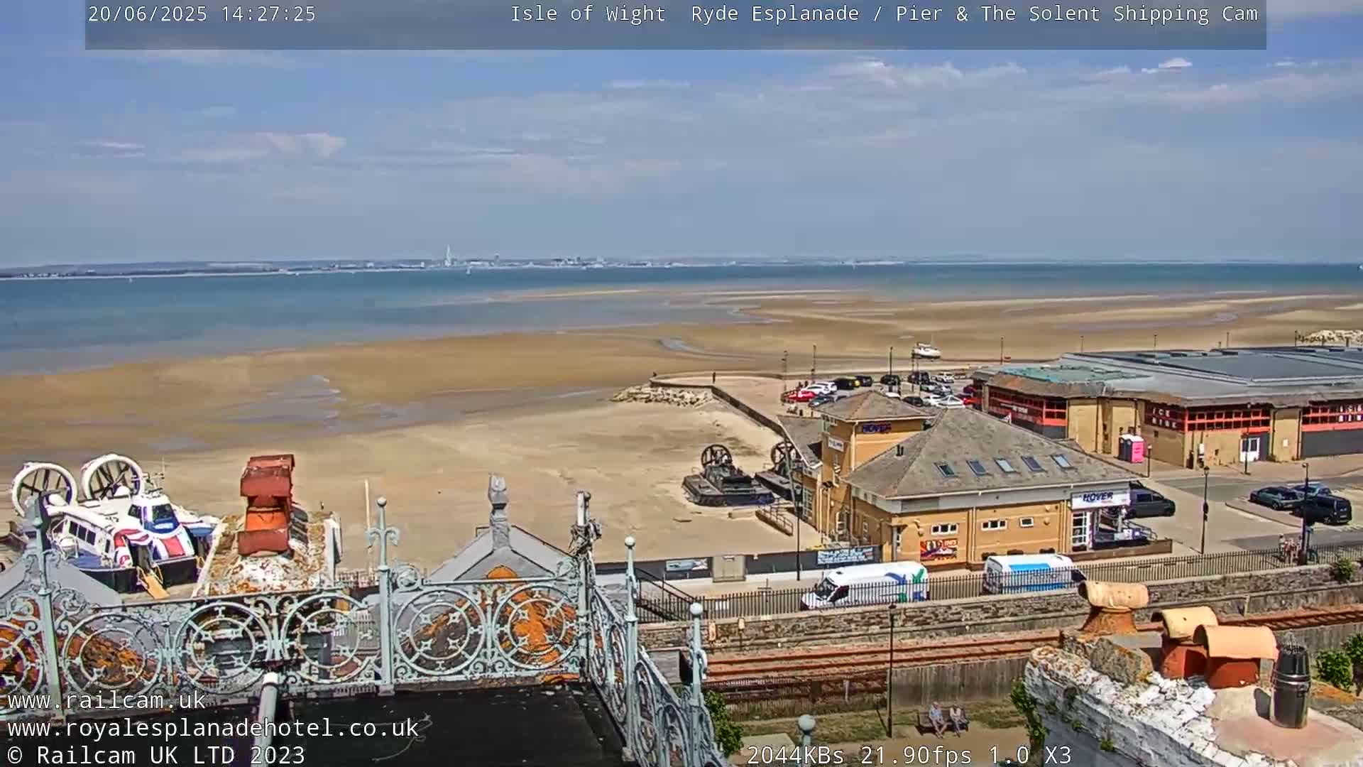 A mostly sunny view overlooking a sandy beach, calm ocean, and buildings near a pier.