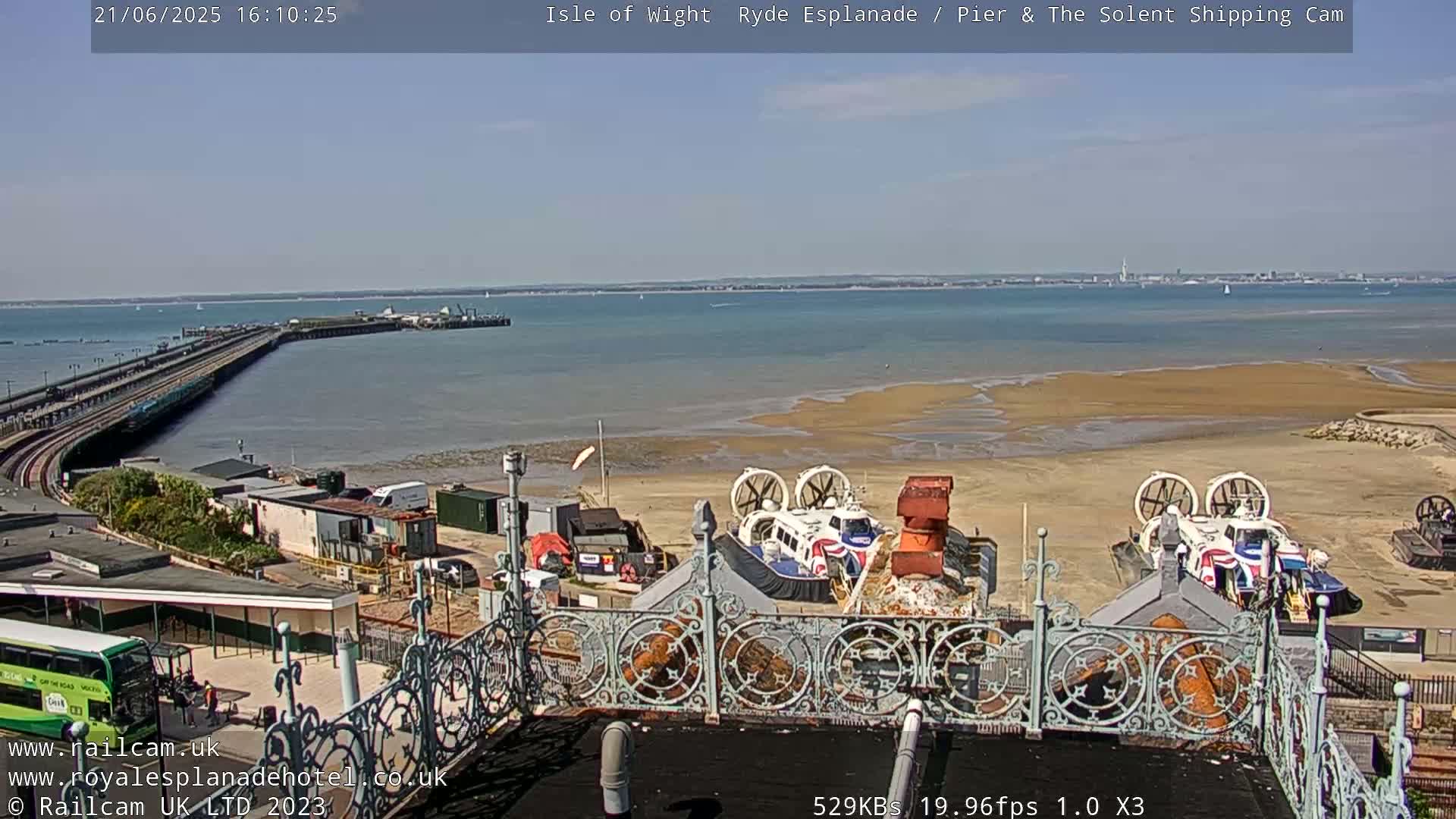 A sunny day overlooking a pier, calm water, a sandy beach with hovercrafts, and a bus.