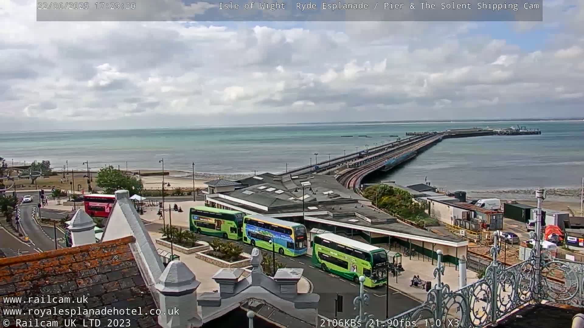 A long pier extends into a calm sea under a partly cloudy sky, with several buses parked near a building at the pier's base and a beach visible in the distance.