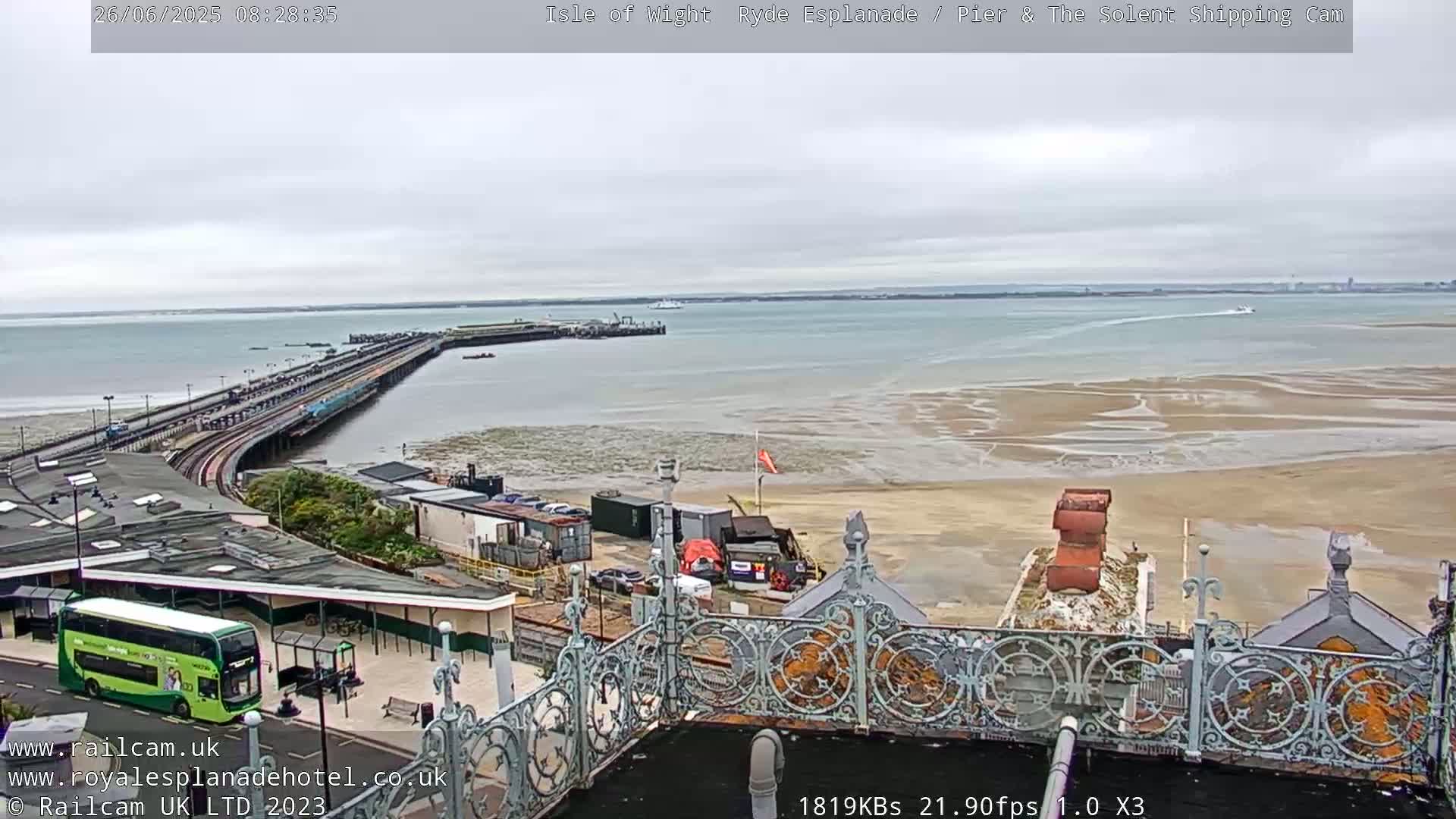 An overcast day shows a long pier extending into a calm body of water with a sandy shore and a bus at a station in the foreground.