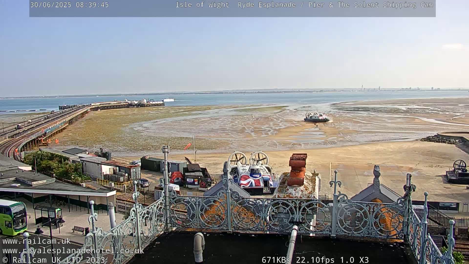 A long pier extends into a wide expanse of shallow, sandy water under a mostly sunny sky, with several hovercrafts visible near the shore.