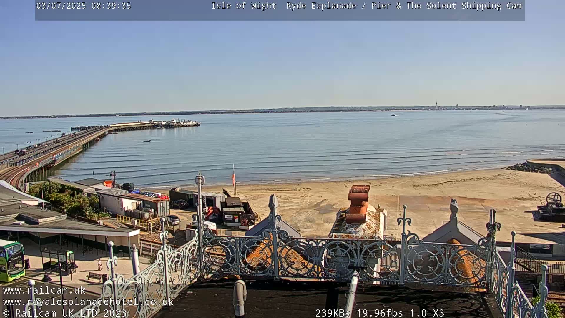 A long pier extends into a calm, sunny ocean with a sandy beach and some construction equipment visible on the shore, seen from an elevated vantage point.